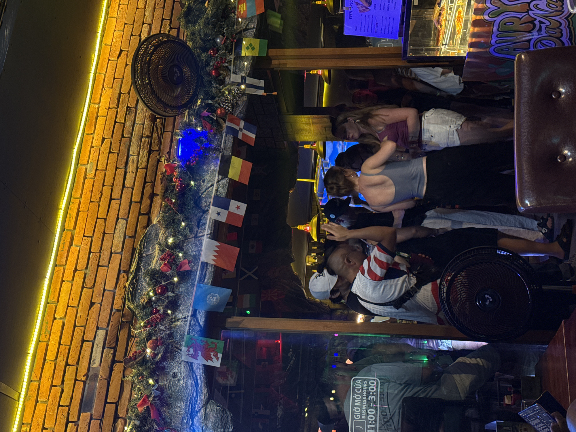 The image depicts the interior of a bar or restaurant in Da Nang, Vietnam, adorned with numerous national flags hanging from the ceiling. A brick wall is visible at the top, illuminated by yellow LED lights. A large black fan is positioned to the right of the frame. Several individuals are present, some standing and interacting, while others appear to be seated. In the background, a display case showcases food items, and a menu board lists "EVENT SPECIALS," "FOOD," and "DRINK" options. A sign with Vietnamese text indicating "BUSINESS HOURS 11:00-3:00" is also visible. The overall atmosphere appears casual and social, with people engaged in conversation and enjoying the ambiance.