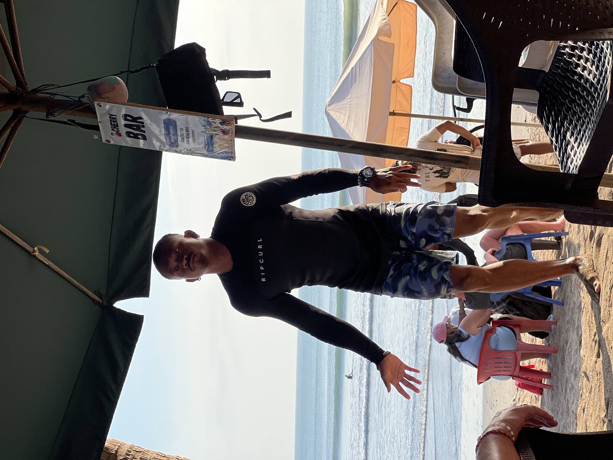 A man in a black long-sleeved rash guard with "RIPCURL" written on it and camouflage shorts stands on a beach in Kuta, Indonesia. He is looking towards the camera with his left arm extended as if beckoning. In the background, the ocean is visible with a surfer in the distance. Several beach chairs and umbrellas are set up on the sand. A sign for "CHERRY BAR" hangs from an umbrella pole. The sky is clear and the sun is shining.