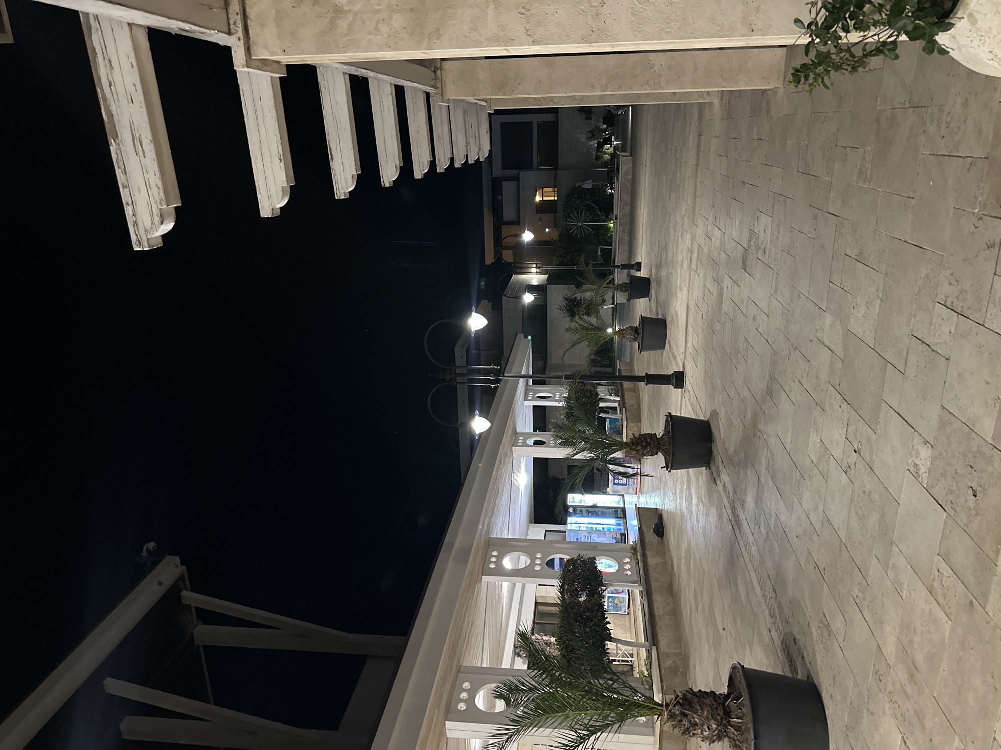 An outdoor walkway at night in Sozopol, Bulgaria, features paved stone flooring and a covered walkway with white beams and support columns. Several street lamps with glowing globes illuminate the area, casting light on potted palm trees and dark-colored planters. In the background, the lights of vending machines are visible within the covered structure. Buildings with illuminated windows are visible further in the distance. The sky is dark, indicating nighttime.