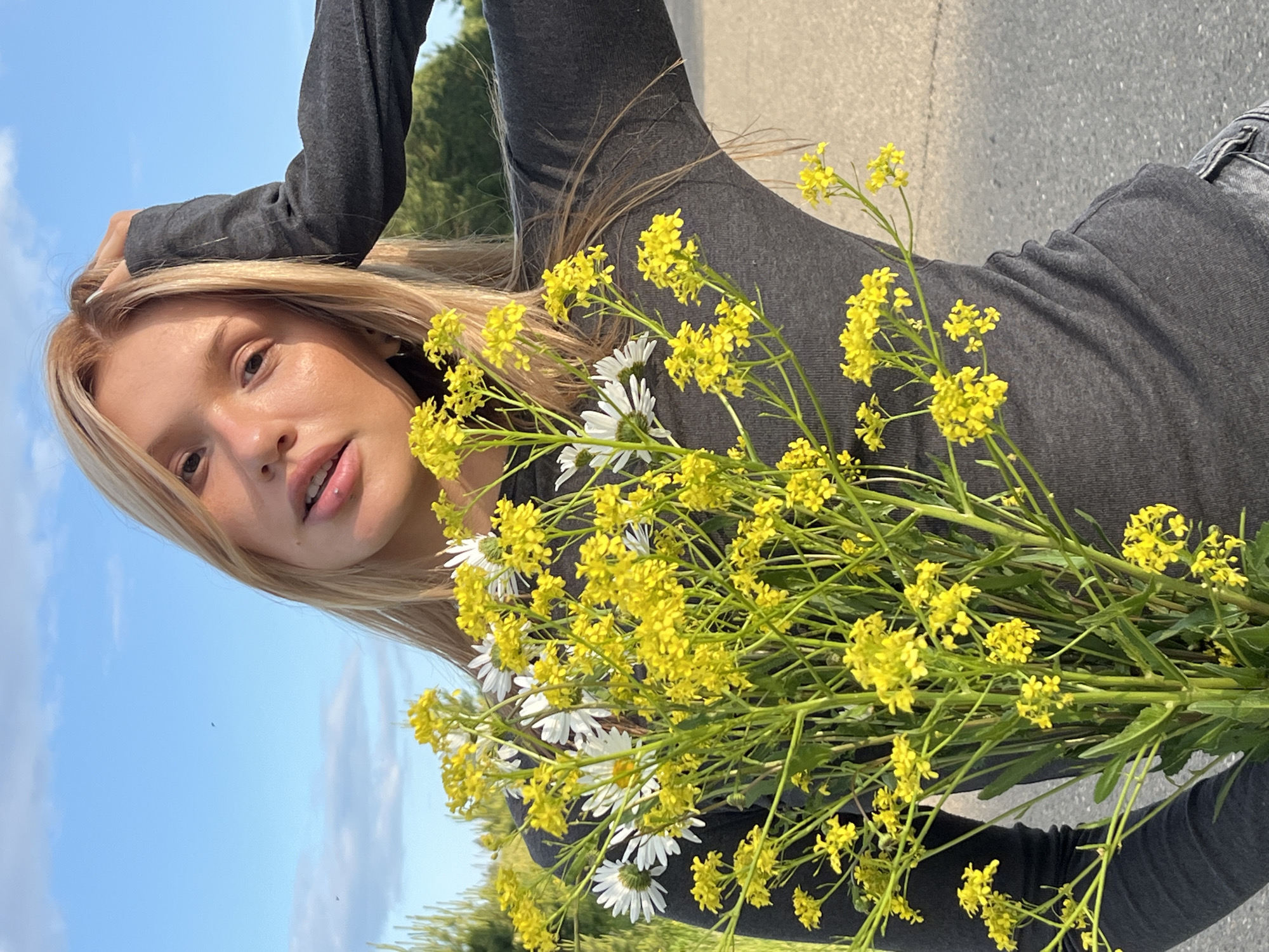 This image captures a young woman posing outdoors on a bright, sunny day, likely in Bol'shoi Bukor, Russia. She is positioned horizontally in the frame, her head towards the left, with long, straight blonde hair flowing around her. She has fair skin, natural makeup, and a serene, direct gaze towards the viewer. A small lip piercing is visible on her upper lip.
The woman is wearing a dark gray, long-sleeved top and what appear to be dark grey jeans. Her left hand is raised to her head, with light-colored nail polish visible. She is holding a substantial bouquet of wildflowers, which are a prominent feature of the image. The bouquet consists primarily of small, numerous vibrant yellow flowers, reminiscent of wild mustard, mixed with several larger white daisies. The flowers partially obscure her body, extending across the center and lower portions of the frame.
The background features a clear blue sky with a few wispy white clouds, indicating fair weather. Below the sky, a gray asphalt road or path is visible, suggesting a roadside or open field setting. Distant green trees and foliage further define the natural, possibly rural or semi-rural, environment. The bright, even lighting indicates it is daytime, likely during the morning or afternoon. There is no visible text in the image.