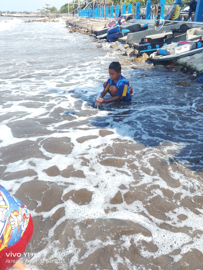 The image depicts an outdoor coastal scene in Limbangan, Indonesia, under bright daylight. A young boy, approximately 6-10 years old, with dark hair and wearing a blue and yellow short-sleeved shirt with a graphic design, is centrally positioned. He is barefoot, crouching in shallow, frothy seawater near the shoreline. The boy is holding a transparent plastic bottle, appearing to interact with the water, possibly filling or pouring it.
The immediate foreground shows brown sandy seabed partially obscured by white seafoam from incoming waves. A portion of a person's leg wearing red shorts with a white and blue astronaut-themed print is visible in the bottom-left corner of the frame.
To the right and in the background, a rocky and concrete embankment defines the edge of the water. Several small, blue-accented boats or canoes are stored on this embankment, some appearing to be overturned. A blue railing or fence runs along the top of the embankment. Beyond this structure, further in the distance, are indistinct buildings or structures and sparse vegetation under a clear, bright sky.