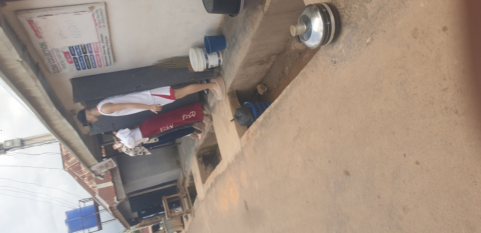 A street scene in Jos, Nigeria, shows a row of shops with open doorways. In front of one shop, a sign reads "GLOW COLLECTION HOME OF THRIFT WEAR." Below this, a list of items for sale is displayed, including "TOPS, GOWNS, TROUSERS, SHIRTS, SKIRTS, AND JACKETS." A handwritten note on the sign says "GIRL NEEDED." Several mannequins are positioned outside the shop, one wearing a white tank top and red shorts, and another wearing a white top and red patterned trousers. Nearby, a blue plastic basket holds a black garbage bag, and several buckets are also visible, one white and one blue. In the foreground, a metallic thermos sits on the ground next to a concrete drainage ditch. The sky is overcast.