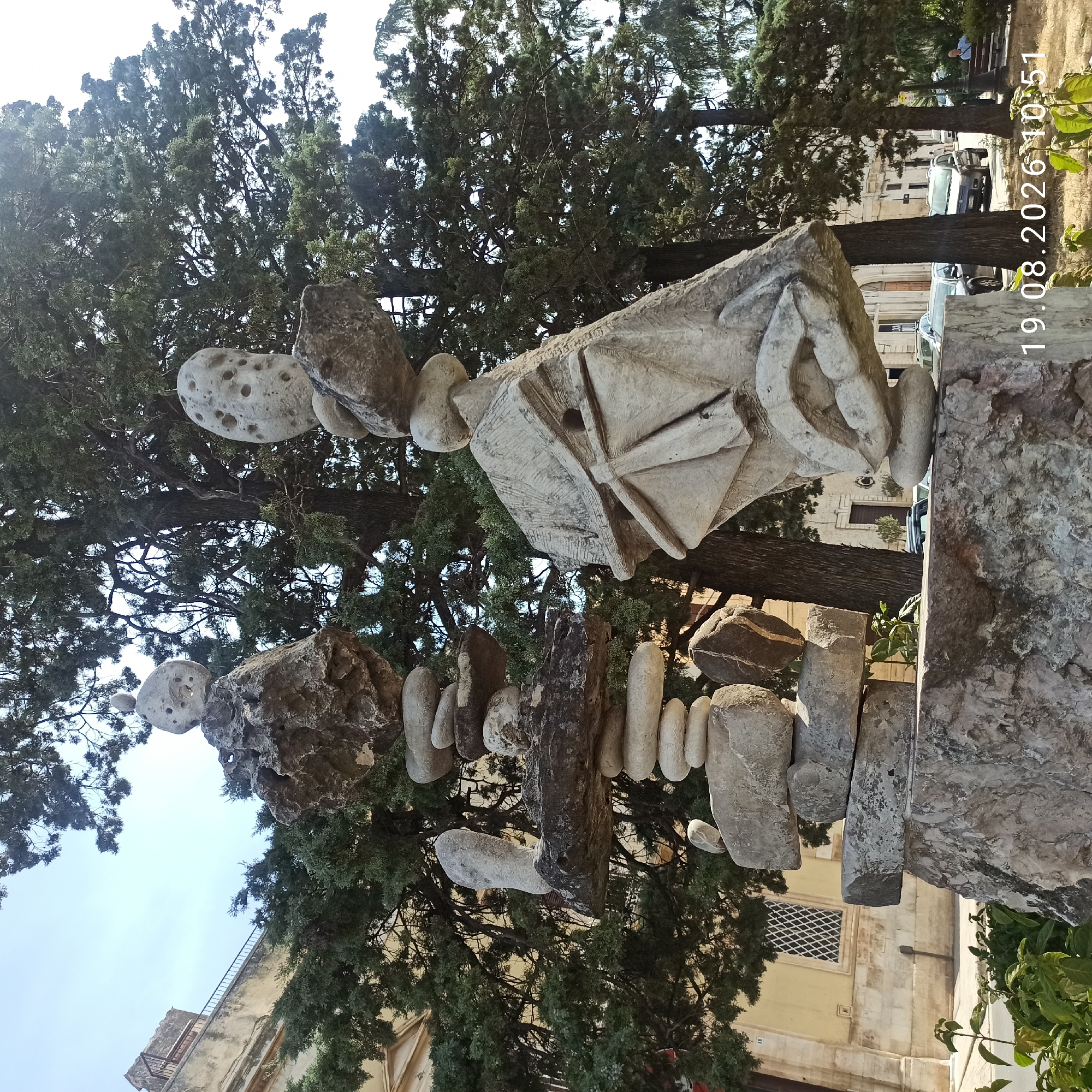Two abstract stone sculptures are prominently displayed in an outdoor setting, possibly a public park or garden, in Ruvo di Puglia, Italy. The sculpture on the left consists of stacked stones of varying shapes and sizes, culminating in a roughly spherical stone with multiple holes. A smaller, elongated stone protrudes horizontally from the middle of this structure. To the right, a larger, more stylized sculpture dominates the frame. This piece features a carved form that resembles a face or mask, with what appear to be lips and a structure reminiscent of a house or building with a peaked roof. It is also constructed from stone and topped with a hole-ridden spherical element, similar to the other sculpture.
Behind the sculptures, a large, mature tree with dense green foliage provides a natural backdrop. The tree's branches extend across a significant portion of the upper frame. In the background, a pale yellow building with architectural details, including a lattice-windowed opening, is visible. A street with parked cars and a few people can be seen beyond the garden area. One individual is seated on a bench to the right. The overall environment suggests a sunny day with clear skies. The bottom right corner of the image displays a timestamp: "19.08.2026 10:51".