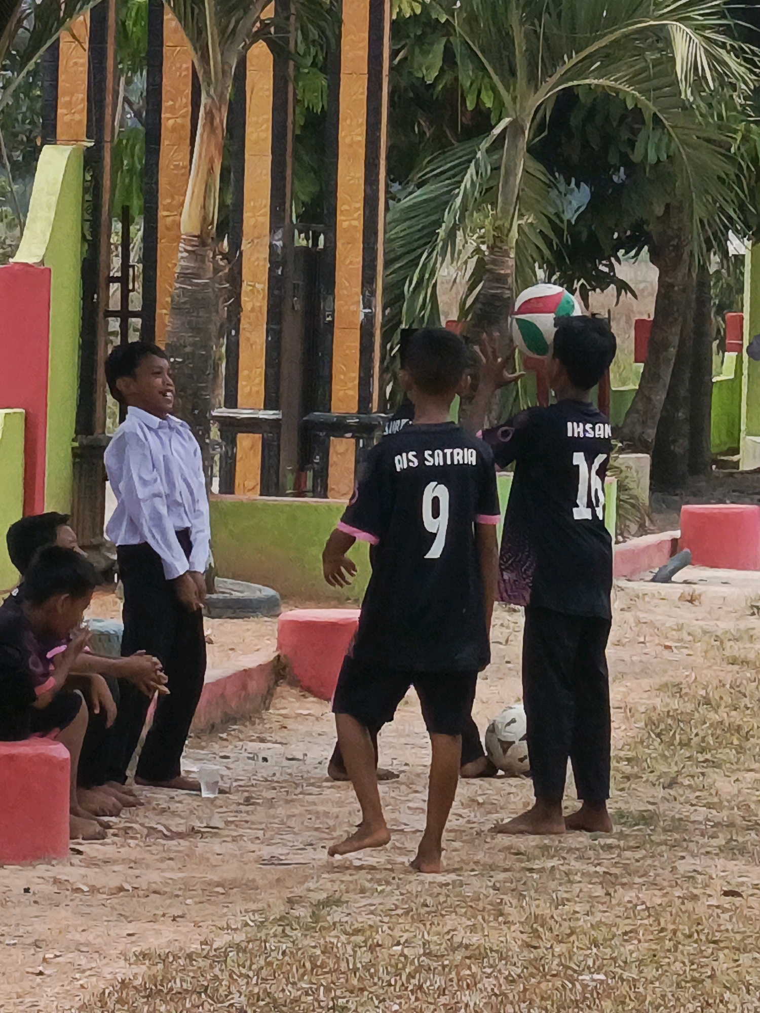 In Megang Sakti V, Indonesia, a group of young boys are gathered outdoors on a dirt and grass area. In the foreground, three boys are visible, two standing and one seated. One standing boy, wearing a light blue dress shirt and black pants, laughs while looking to his right. Another standing boy in a black shirt with the name "IHSAN" and the number "16" on the back, is in mid-action, appearing to toss a volleyball. To his left, another boy wears a black shirt with "AIS SATRIA" and the number "9" on the back. In the midground, a volleyball is in the air, and a soccer ball rests on the ground. The background features a fence with vertical orange and black stripes, green foliage, and palm trees. Red and green structures are present on the left side of the image, and red cylindrical objects are visible in the right midground.