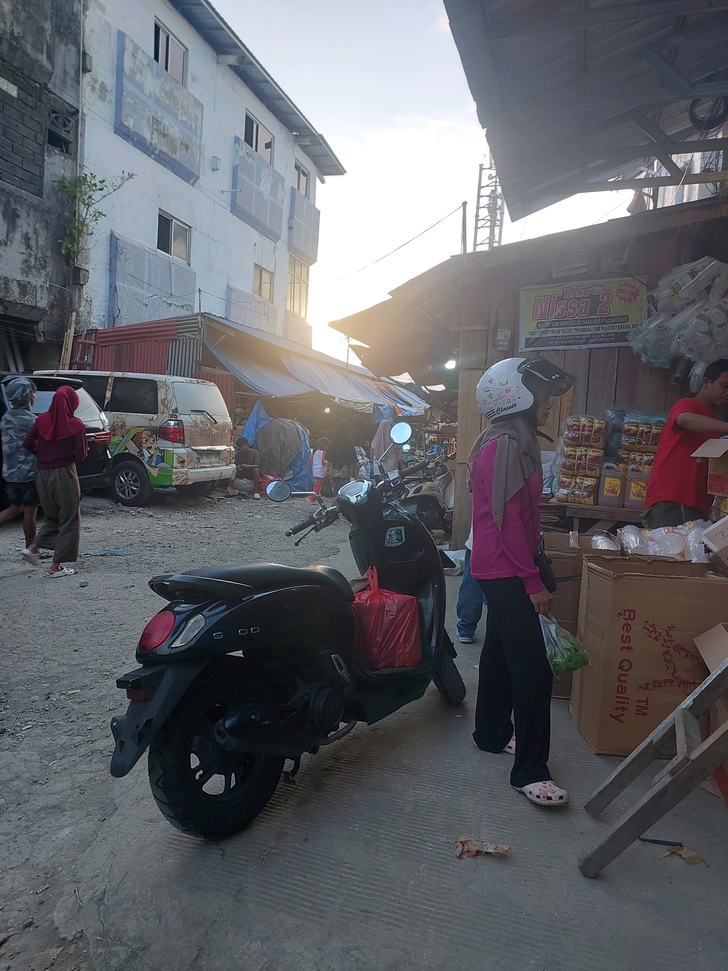 In Ambon, Indonesia, a street scene unfolds in what appears to be a market area. The sun casts a warm glow, highlighting the weathered facade of a multi-story building on the left, with visible signs of age and some graffiti. In the foreground, a black scooter is parked, its rear wheel and taillight clearly visible. A woman in a pink shirt, grey headscarf, and dark pants, wearing pink clogs, stands near the scooter, holding a plastic bag of green vegetables. A red plastic bag is attached to the scooter's rear.
Further into the scene, another scooter is parked. A van, adorned with cartoon characters, is parked to the left of the scooters. In the background, a man in a red shirt is interacting with a stall that displays various packaged goods, including what appear to be instant noodle packs. Above the stall, a banner reads "Toko Nissa 2 Melayani Berbagai Macam Packaging". The environment suggests a bustling marketplace, with awnings and makeshift structures indicating commercial activity. The ground is paved with concrete and dirt.