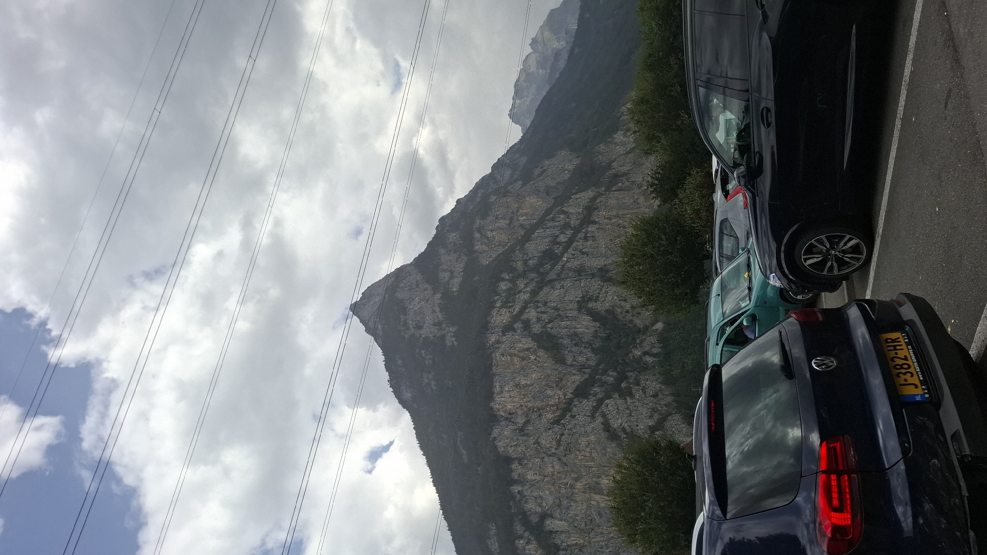 In Erstfeld, Switzerland, a mountainous landscape dominates the background of this image. A prominent, craggy peak rises sharply, its slopes covered in dark, dense trees at the lower elevations and exposed rock higher up. The sky is overcast with dramatic, grey clouds, punctuated by patches of blue, and several power lines crisscross the scene horizontally in the upper portion. In the foreground, a parking lot is visible, occupied by several vehicles. A dark blue SUV is parked on the left, facing away from the camera, with its taillights illuminated. A bright yellow license plate with black lettering "J-382-HR" is affixed to its rear. To the right of the blue SUV is a black SUV, and further back, a teal-colored van is parked, with a person's head visible through the driver's side window. The parking lot is paved and lined with white markings. Trees line the edge of the parking lot between the vehicles and the mountain.