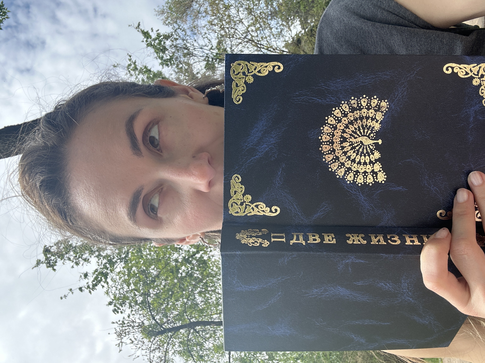 A woman is holding a book titled "Две Жизни" (Two Lives) in front of her face, possibly while sitting outdoors. The book has a dark blue cover with gold embellishments, including a peacock design and corner decorations. The woman has light skin, brown eyes, and her hair is pulled back. The background shows a cloudy sky and green foliage, suggesting a park or other natural setting. Without more information or coordinates, pinpointing the specific location in Kryvyi Rih is not possible.