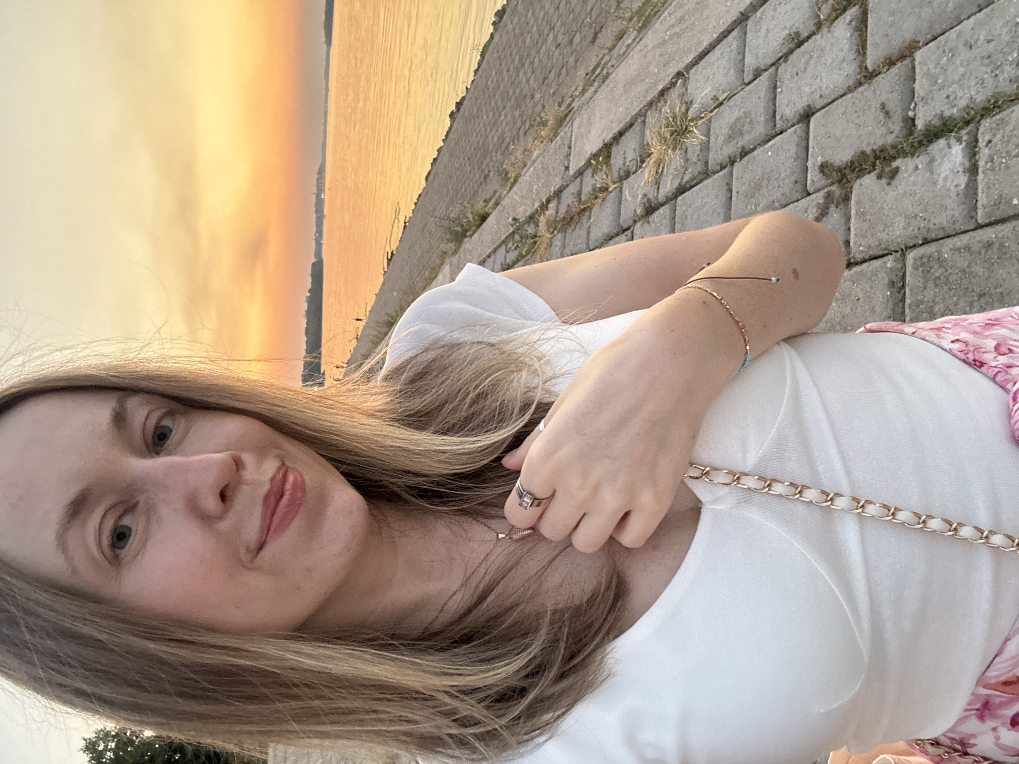 A woman with blonde hair takes a selfie outdoors with a river and a sunset in the background. She is wearing a white top and a pink floral skirt. She has jewelry on her neck, wrist, and fingers. The paved walkway she is standing on is adjacent to the river bank. The scene appears to be a river promenade in Belgrade, Serbia, likely near the Sava or Danube River, during a sunset.