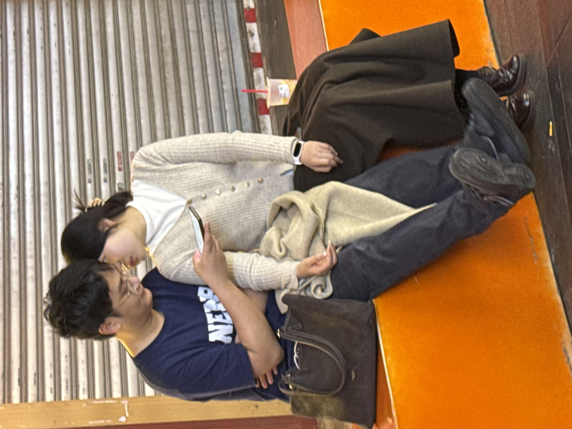 A couple is seated on an orange bench in front of a closed, corrugated metal storefront. The man, wearing glasses and a dark blue t-shirt, has his arm around the woman, who is looking at a cell phone. The woman wears a light-colored cardigan and dark pants, and a beige scarf is draped over her legs. A dark brown purse sits on the bench beside the man. A beverage with a red straw sits on the ground nearby. It is not possible to verify the specific location in the city of Liangjiang, China, using only the information contained in the picture.