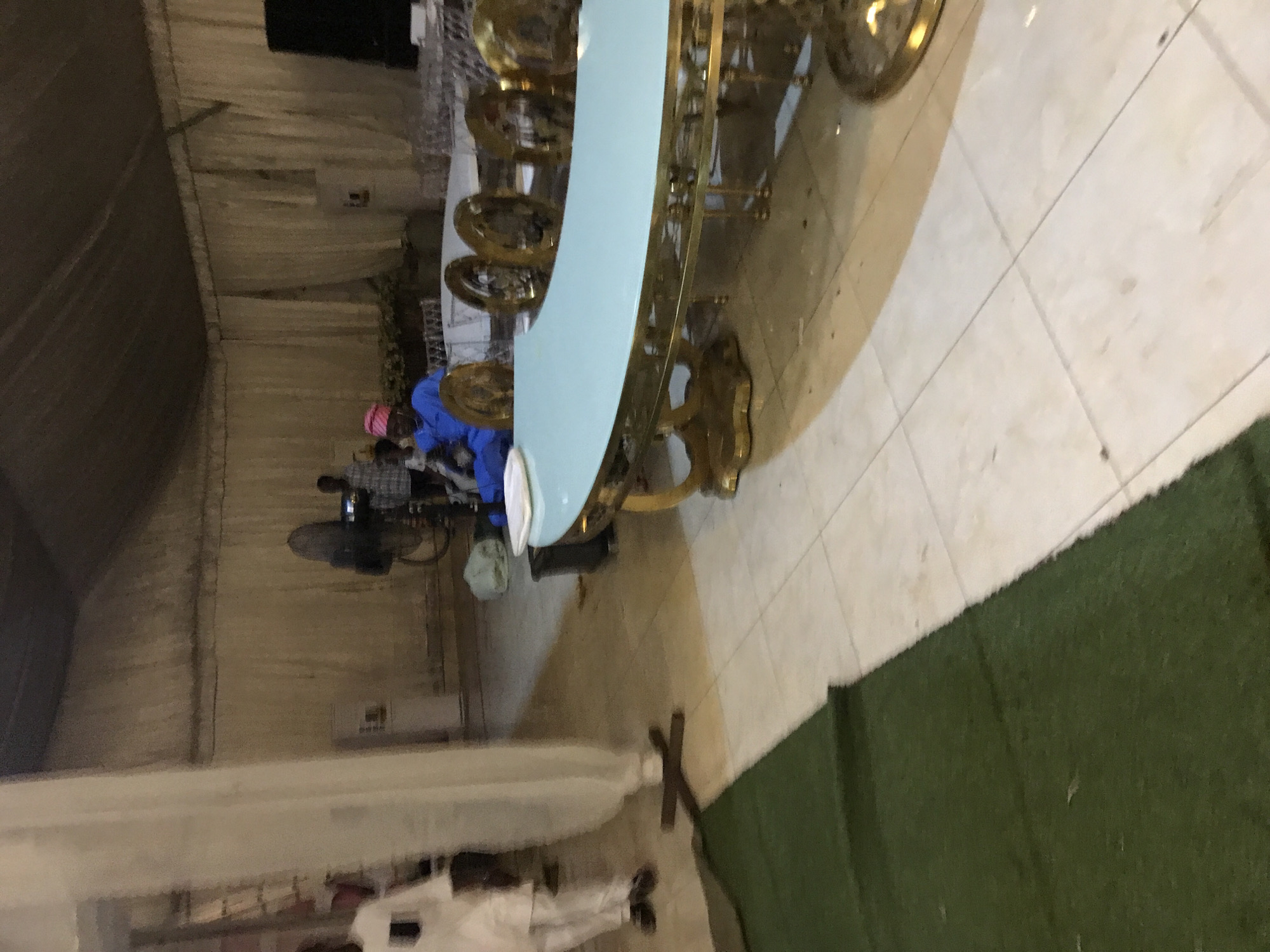 The image shows an indoor setting, likely a hall, in Oko Erin, Nigeria. A large, light blue curved table with gold ornate legs and multiple chairs around it is prominently displayed. People are present, including individuals dressed in blue and pink attire near the table, and others in white garments towards the bottom of the frame. A black standing fan is also visible. The flooring is a mix of light-colored tiles and a strip of green artificial turf. The walls are draped with fabric, suggesting the space is set up for an event.