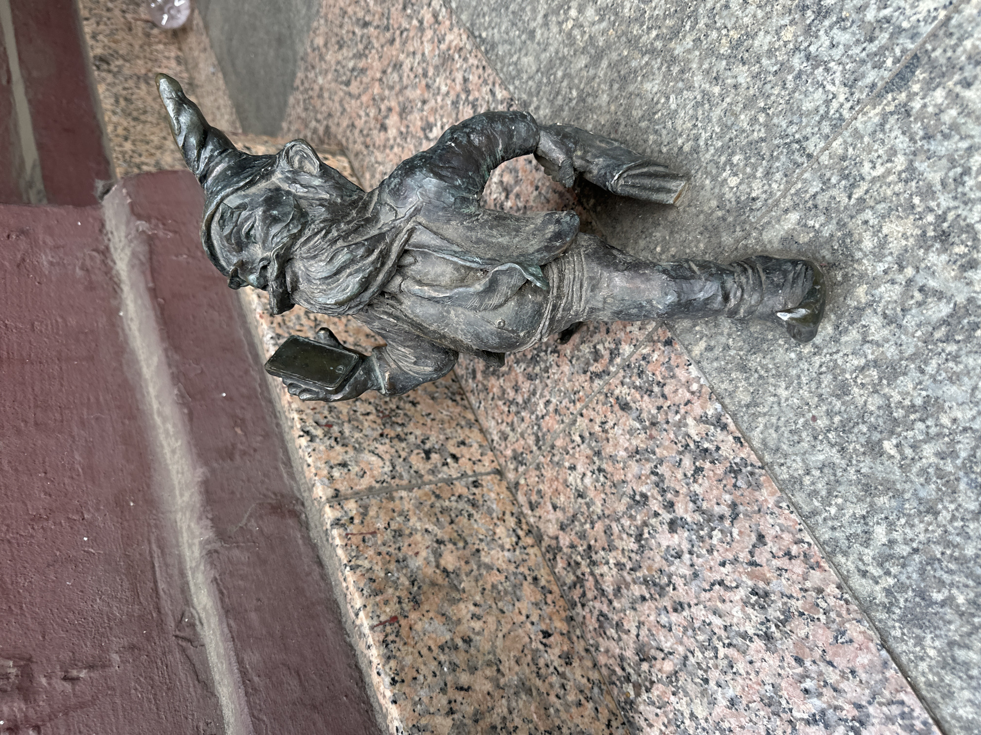 A bronze dwarf statue is on a stone surface in Wrocław, Poland. The dwarf wears a pointed hat and holds a rectangular object resembling a smartphone. The statue's placement is next to a set of steps, with the stone surface transitioning between granite and a smoother gray stone.