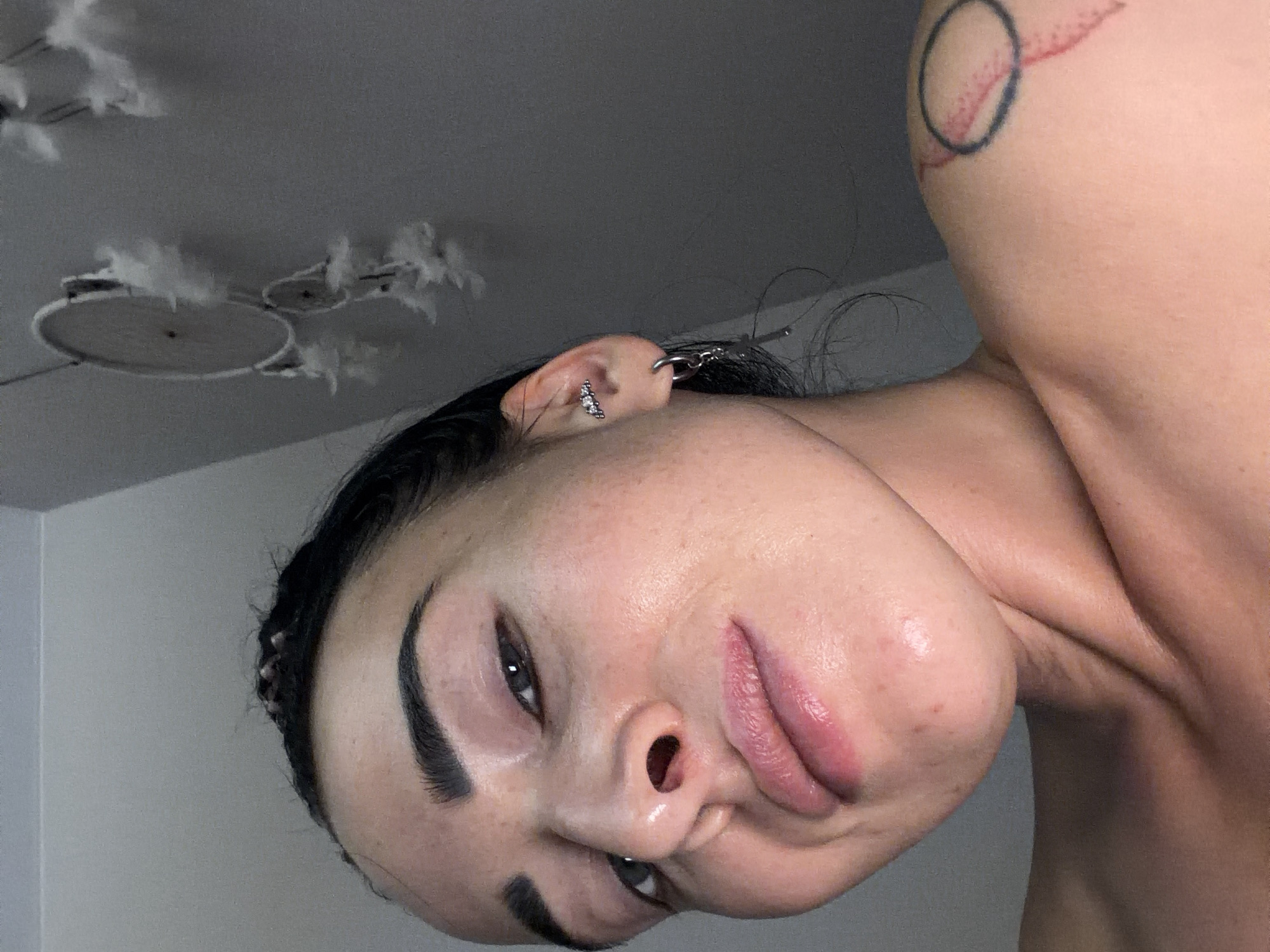 The image features a close-up portrait of a person, likely female, whose head is tilted significantly to the left, almost appearing horizontal. The person has a clear complexion with a natural, dewy finish, dark, well-defined eyebrows, and dark eyes looking towards the viewer. Their full lips are slightly parted, showcasing a soft, pink hue.
Multiple piercings are visible on the left ear, including a cluster of small gems on the lobe and a large silver hoop with a dangling charm (possibly a key shape) on the upper cartilage. Dark hair is pulled back, with some loose strands visible around the ear. On the person's right shoulder/upper arm, a tattoo is visible, depicting a circular design in red and black ink.
The background shows a plain, light-colored ceiling. An unusual light fixture or ceiling decoration is visible in the upper left corner of the frame, adorned with several small, white, feathery or fluffy elements. The lighting is soft and even, highlighting the person's features without harsh shadows. No text is visible in the image. The overall impression is an intimate, somewhat casual portrait with a slightly artistic and unconventional composition due to the tilted angle.