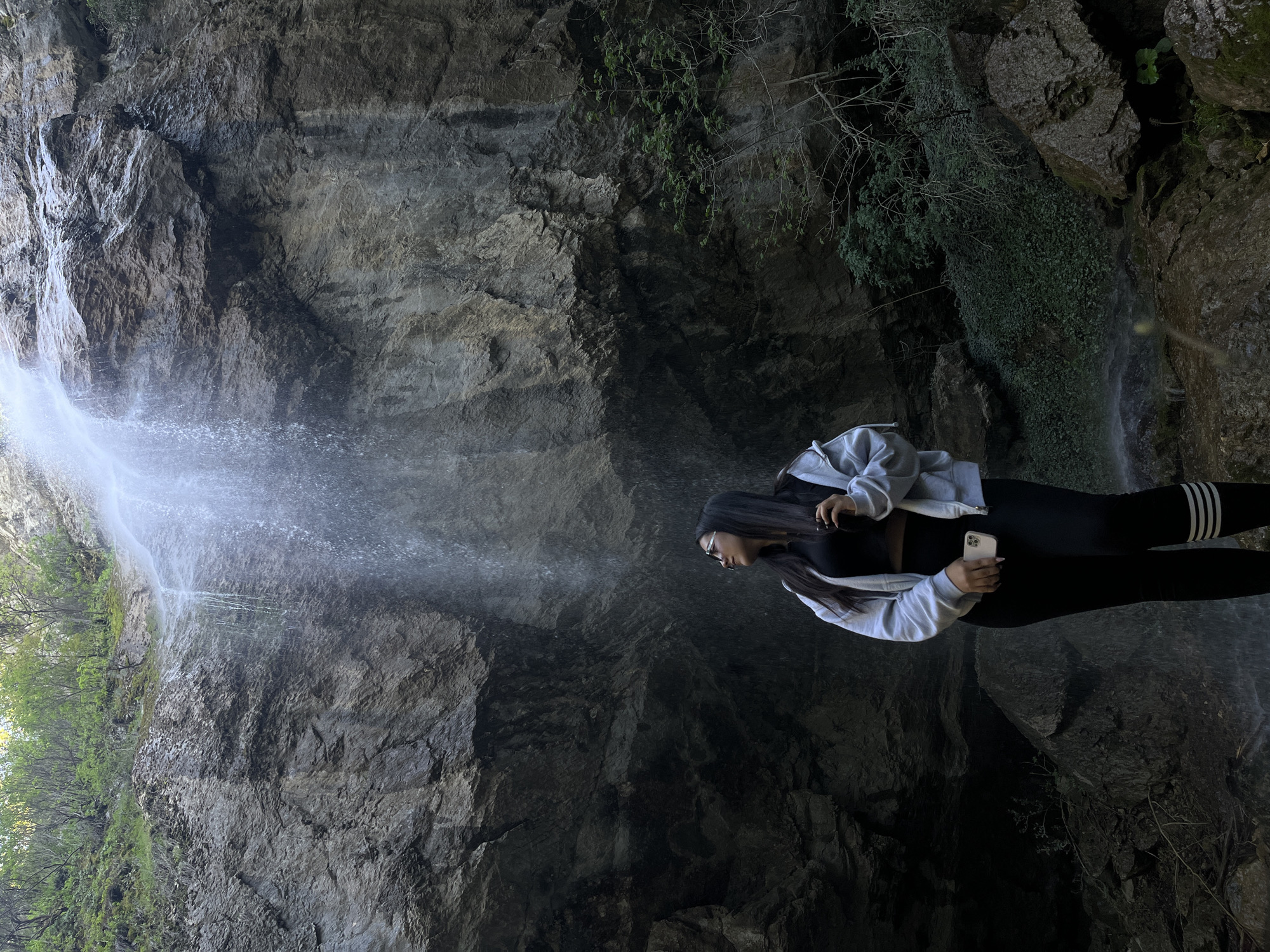 A long-haired woman wearing a black top and pants with white stripes down the side, a light gray zip-up jacket, and glasses stands holding a phone near a waterfall. The location is Aleksandrovo, Bulgaria, near a rocky, cliffside landscape with lush greenery. The waterfall cascades down a rocky wall. The woman is positioned on the right side of the image, while the waterfall and rock face dominate the left.