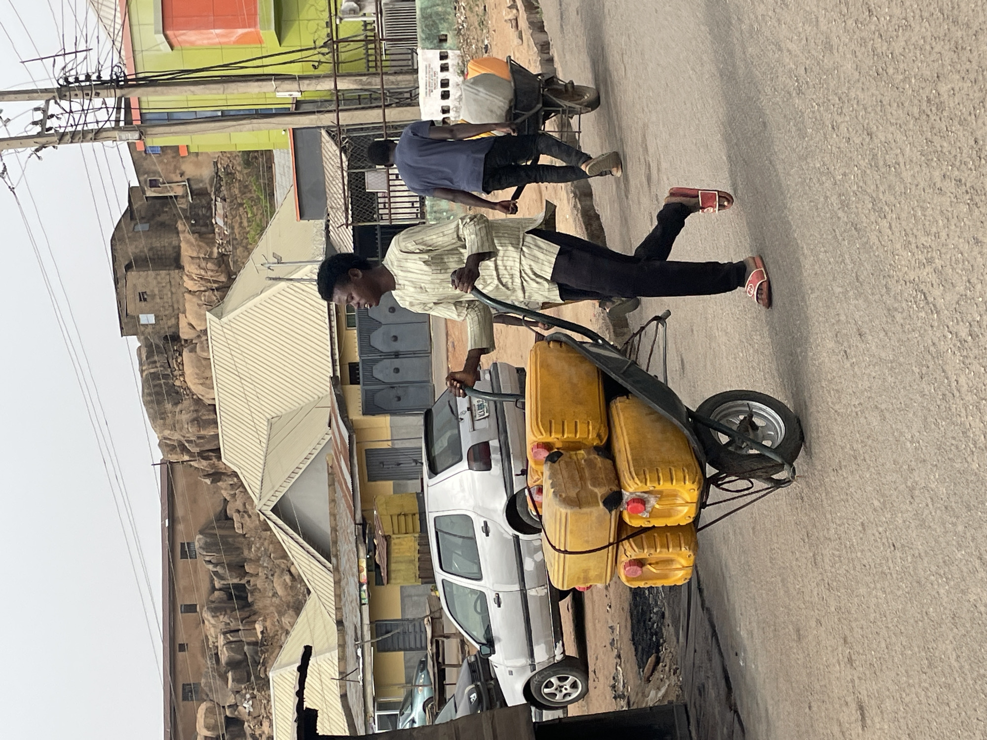 A man wearing a striped shirt and black pants is running on a paved street, pulling a wheelbarrow loaded with multiple yellow jerrycans. He's wearing red sandals. Another man in a blue shirt and black pants is walking nearby. The scene is set in a city, with buildings of various heights and materials visible in the background, including a building painted in green and orange, and others with corrugated metal roofs. Cars are parked alongside the road. The presence of electrical wires suggests an urban setting. The background also shows rock formations. Based on this, it appears that the photo was taken in Jos, Nigeria.
