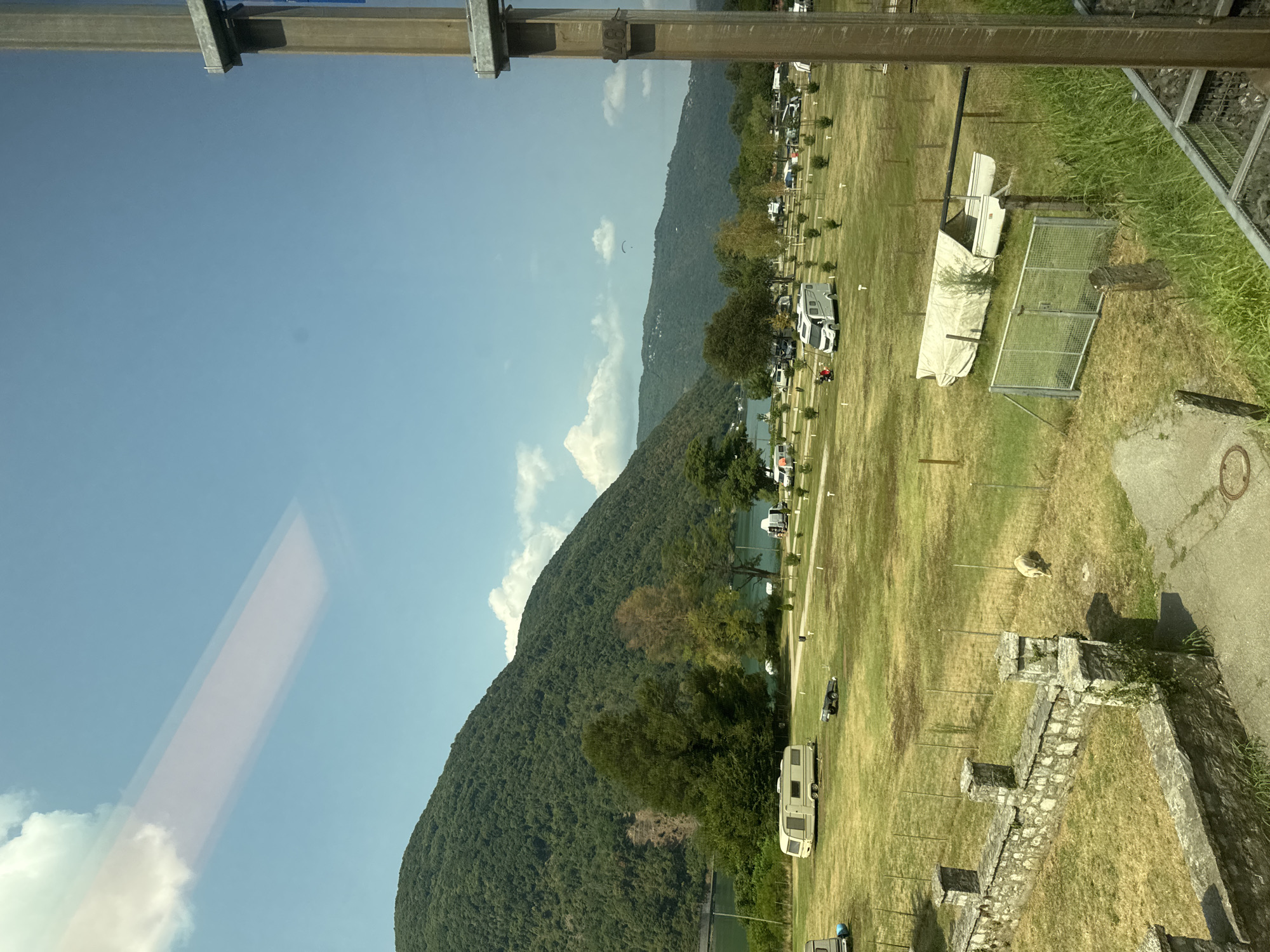 The image depicts a view from a window, likely a train, looking out at a waterside campground. A large, tree-covered mountain dominates the left side of the frame, with a body of green water visible at its base. The campground is spread across a grassy field, populated with several caravans, RVs, and what appears to be a covered boat. In the foreground, a low stone wall runs along the edge of a pathway. The sky is blue with a few scattered clouds, and a paraglider is visible in the distance. The scene suggests a tranquil, outdoor recreational area in the city of Val Mara, Switzerland, a location known for its scenic beauty and proximity to Lake Lugano.