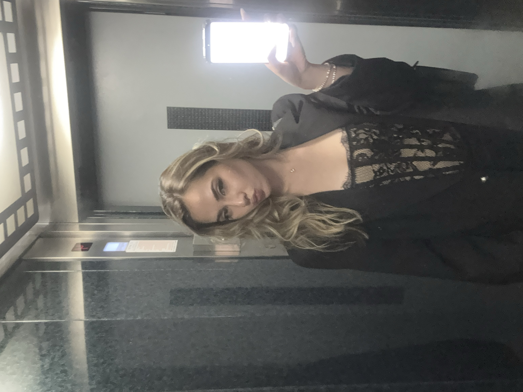 A young woman is taking a selfie in an elevator. She is holding a phone in her right hand, which is illuminating her face. She is wearing a black blazer over a lacy black top. She has long blonde hair. The elevator appears to be a standard model, with metal doors and a panel of buttons. There are no clear indicators of the exact location in Blagoevgrad, Bulgaria.