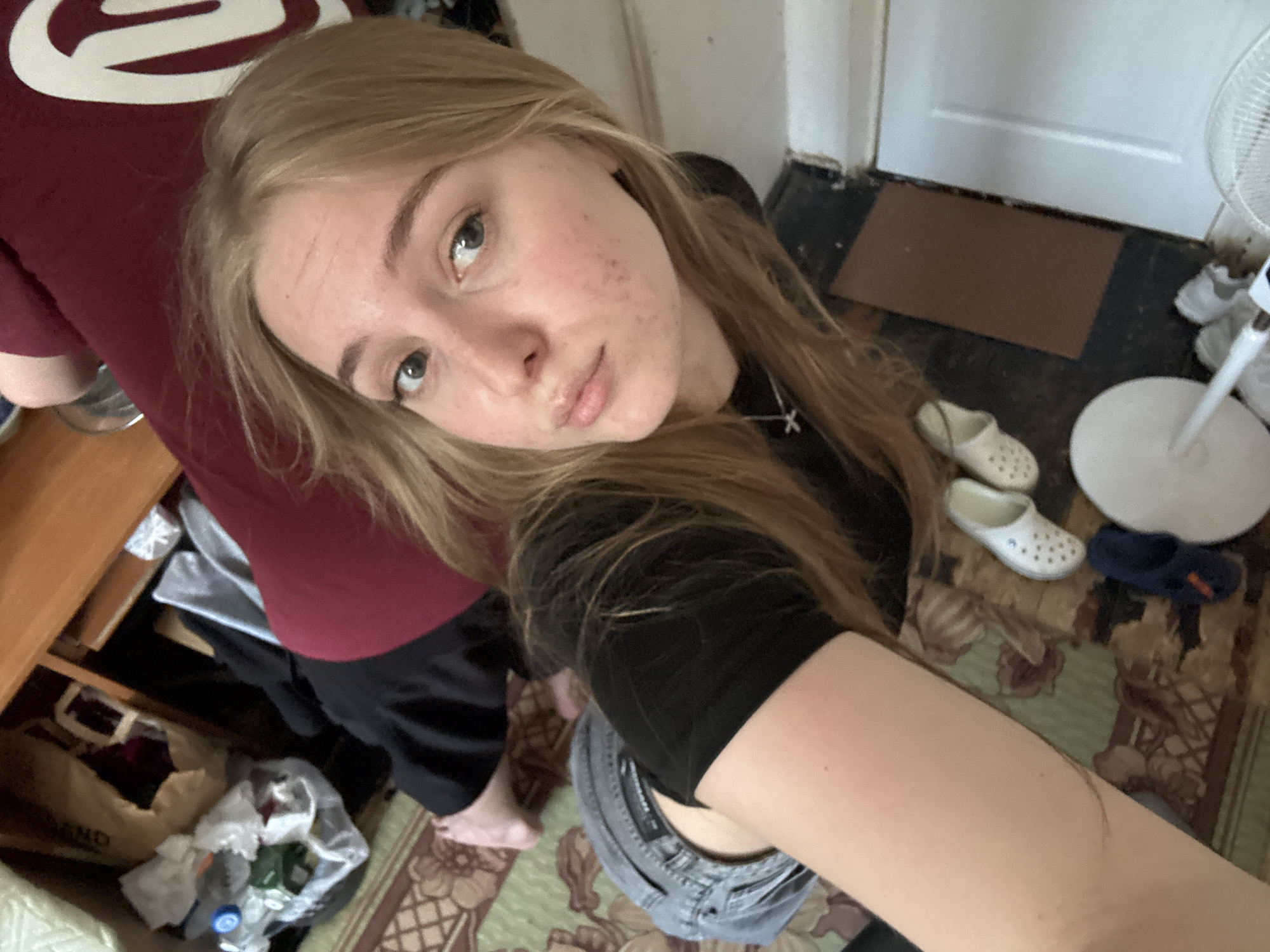 A young woman with blonde hair and fair skin is taking a selfie. She is wearing a black top and a silver necklace with a cross pendant. She is standing next to a person in a maroon shirt. The environment includes a cluttered room with a cabinet, a rug with a floral pattern, a door, a fan, and several pairs of shoes. There is no definitive information to establish the location of this image being in the city of Perm, Russia, or otherwise.