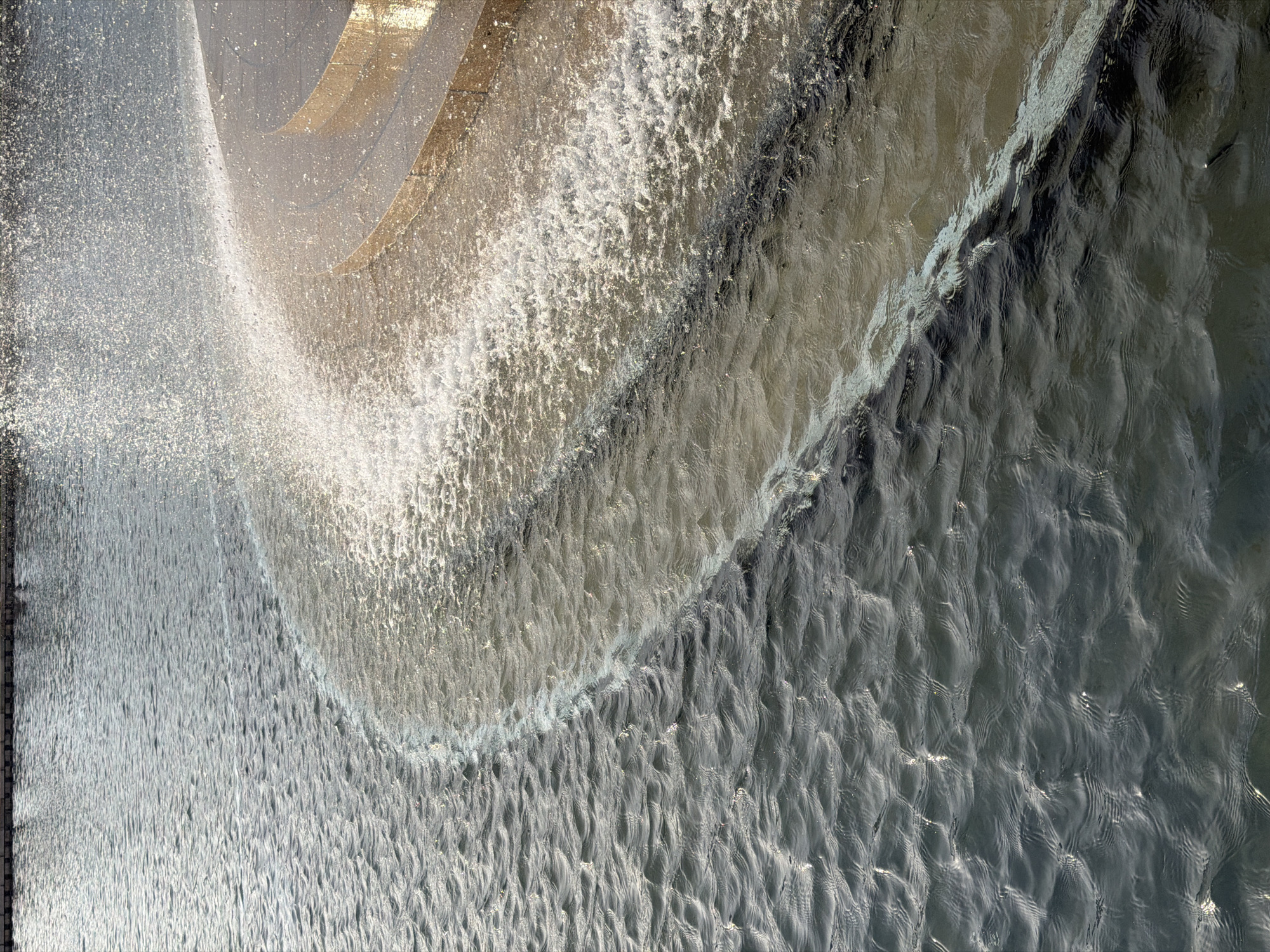 The image is a high-angle shot of a fountain in the city of Krasnodar, Russia. The fountain features a curved, elevated concrete structure from which water cascades down. The falling water creates a curtain-like effect with a visible mist or spray. The water below the cascade exhibits ripples and wave patterns. A metallic grating or railing is partially visible on the left side of the frame. The general tone is silver, gray, and white, due to the water and concrete.