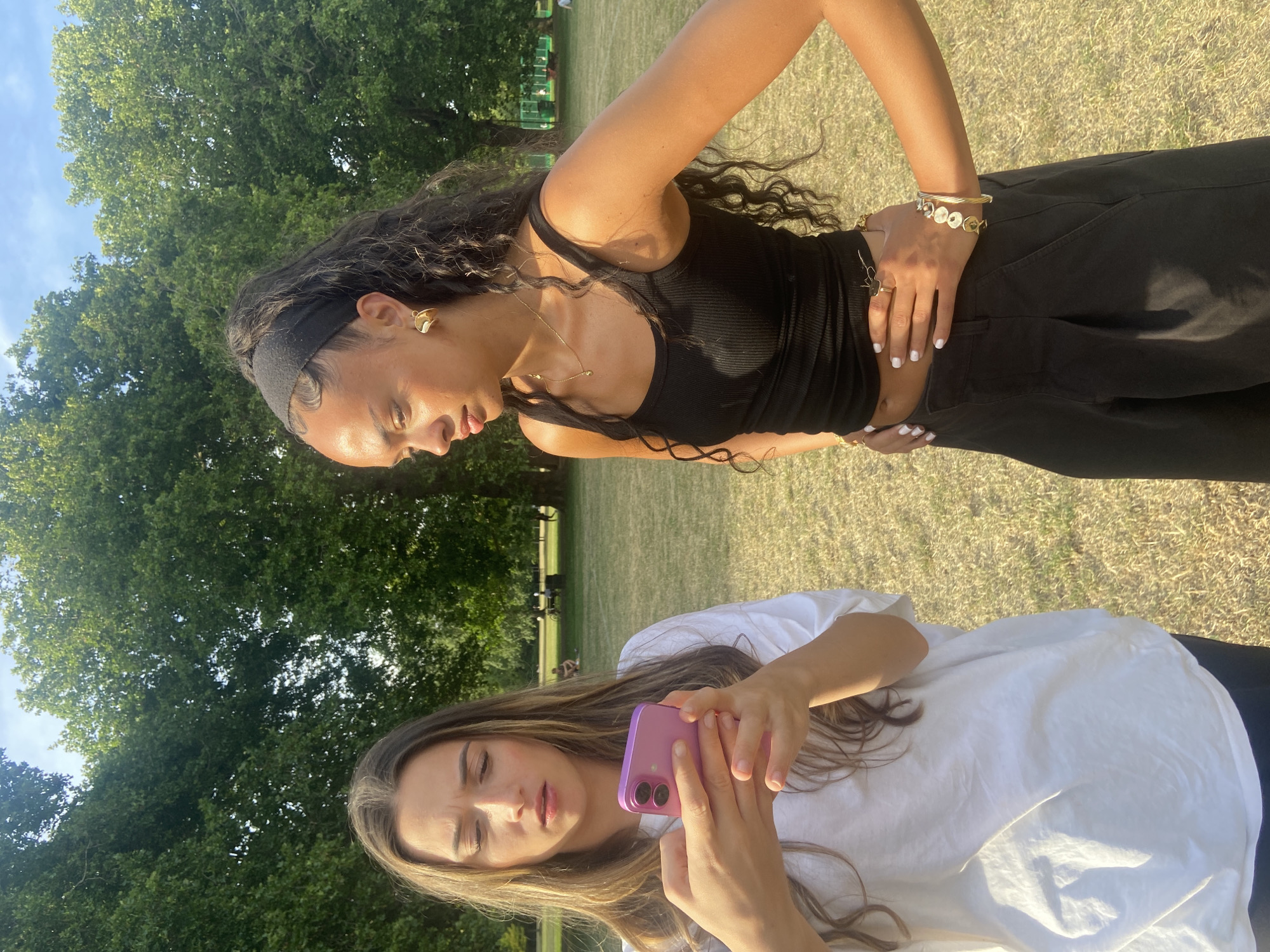 Two young women are standing in a park, likely in London given the presence of mature trees and manicured lawns characteristic of London's Royal Parks or similar city green spaces. The woman on the left, with long, light brown hair, is looking down at a pink smartphone she holds in her hands. Her expression suggests concern or confusion. She is wearing a white t-shirt. The woman on the right, with dark, curly hair pulled back with a black headband, is looking towards the woman with the phone. She is wearing a black tank top and black wide-legged pants, adorned with several bracelets and rings. Her posture is relaxed, with one hand on her hip. The background shows a grassy field with scattered trees and a few distant figures, suggesting a public park on a sunny day.