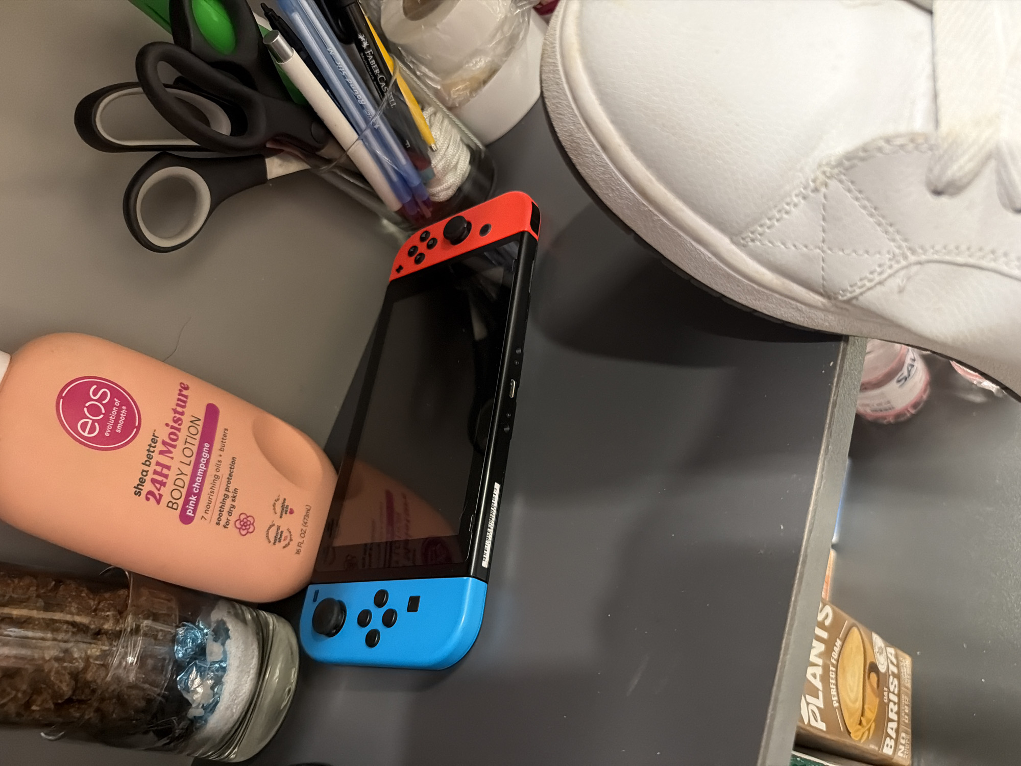 This media file shows a close-up overhead view of various objects on a flat, gray surface, likely a desk or countertop. The arrangement suggests a collection of personal items.
Prominently featured is a Nintendo Switch gaming console with bright red Joy-Cons. To the left of the Switch is a large, peach-colored bottle of EOS Shea Better 24H Moisture Body Lotion in the "pink champagne" scent. Below the lotion bottle is a glass container filled with what appears to be granola or some type of foodstuff, with blue and white material visible at the bottom. In the upper left quadrant, a pair of black-handled scissors is visible, along with a collection of pens and markers in a cylindrical holder, including a white pencil and blue and black pens with visible text like "Faber-Castell" and "Round Stic M." In the upper right, a portion of a white sneaker is visible. A small cylindrical object with red and white labeling is also partially visible. Further to the right, a box with the text "PLANIS PERFECT FOAM OAT BARISTA" is partially visible, suggesting a beverage product.
The lighting is bright and appears to be artificial, casting minimal shadows, suggesting an indoor setting. The overall impression is one of everyday items gathered together, possibly in a home or office environment. No people are visible. The visible text includes brand names and product descriptions. The setting does not provide strong cues for a specific time of day or weather conditions, but the organized yet eclectic arrangement of items implies a personal space.