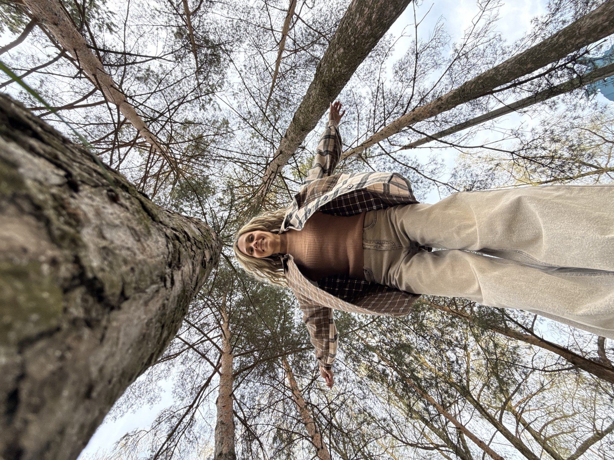 A low-angle shot from the ground level looking up features a young woman as the primary subject. The photo seems to be taken in a forest or wooded area, identifiable by the numerous tall trees surrounding her. The woman is positioned in the middle of the frame, appearing to stand or possibly lie down directly above the camera. She is wearing a brown turtleneck sweater and beige pants, complemented by a plaid shirt. The trees are dense, their branches reaching towards the sky, partially obscuring the light and creating a dynamic perspective. The photo suggests a natural environment, possibly a park, though specific landmarks to confirm the location in Rusanovo, Russia are not visible. The focus is on the subject and the surrounding natural environment, with the camera angle providing a unique view.