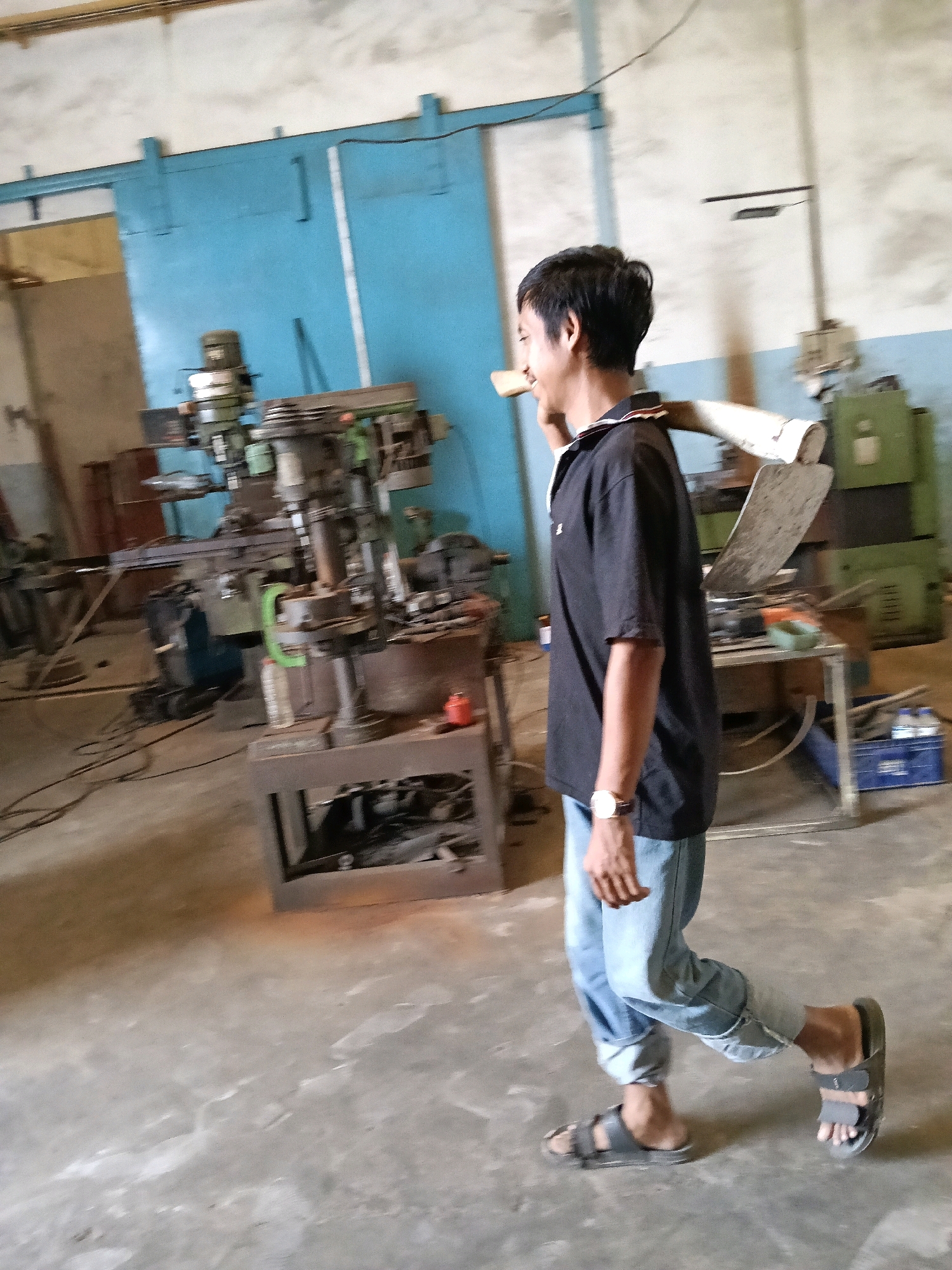 A young Indonesian man with short black hair walks from right to left in an industrial workshop, carrying a large, rolled-up white sheet or canvas over his right shoulder. He is wearing a black polo shirt, light blue jeans rolled up at the ankles, and dark grey slide sandals. A wristwatch with a brown leather band is visible on his left wrist. The workshop appears to be located in Belimbing, Indonesia, and is filled with various machinery and tools, including a milling machine and a drill press. The background features large blue sliding doors and other equipment, suggesting an active production or repair facility. The floor is concrete, with some oil stains and debris. The overall lighting is somewhat dim, with a mix of natural light and overhead artificial lighting. The image is slightly blurred, indicating movement.
