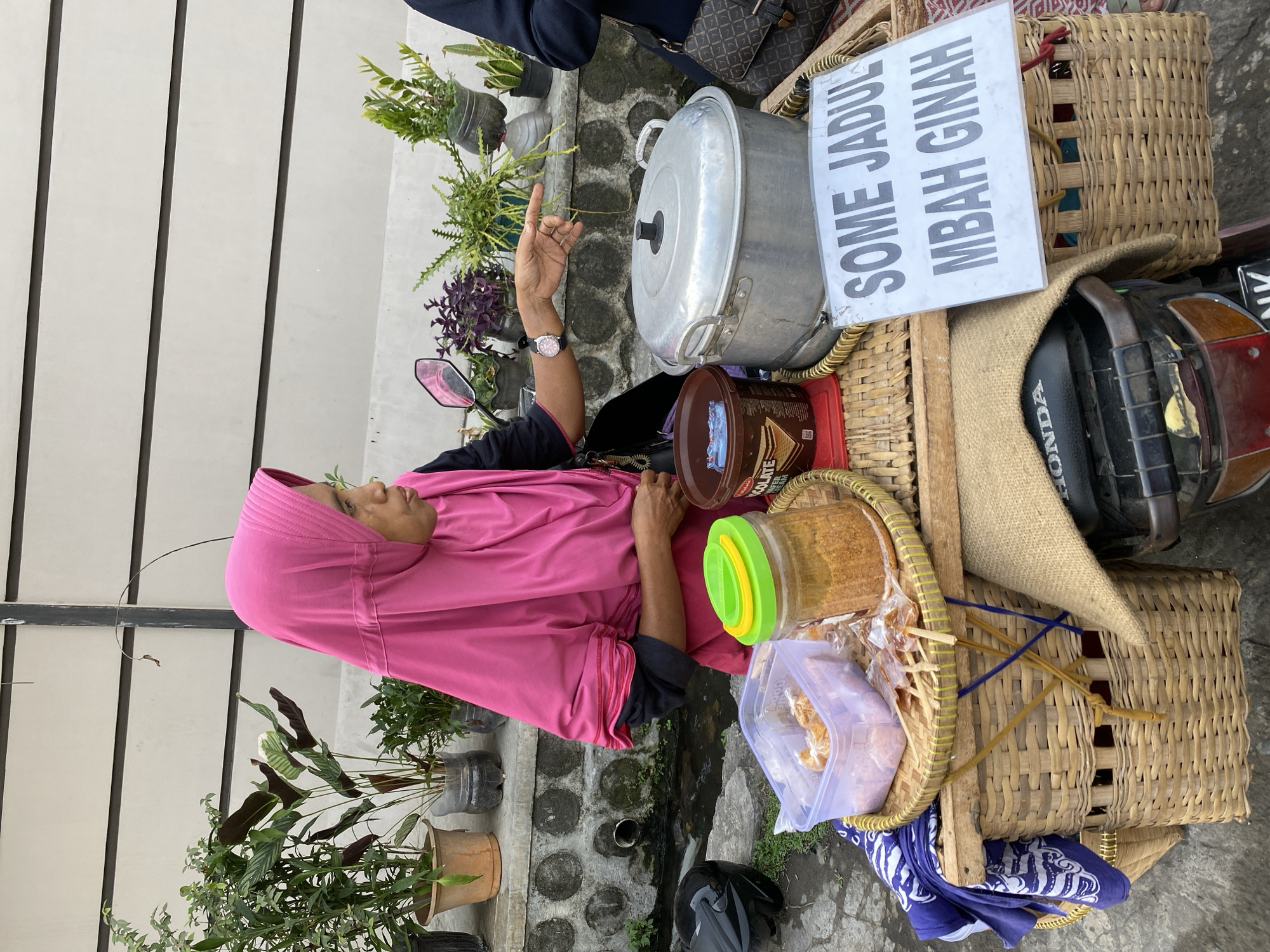 In the city of Kasihan, Indonesia, a woman wearing a bright pink hijab stands behind a food cart. The cart is made of woven bamboo and is attached to a motorcycle. On the cart, there are several items including a large silver pot, a container of food, and a jar filled with an orange, granular substance. A white sign on the cart reads "SOME JADUL MBAH GINAH". Behind the cart, there are potted plants on a low wall, and the ground appears to be paved. The woman is looking to her right and pointing with her left hand.