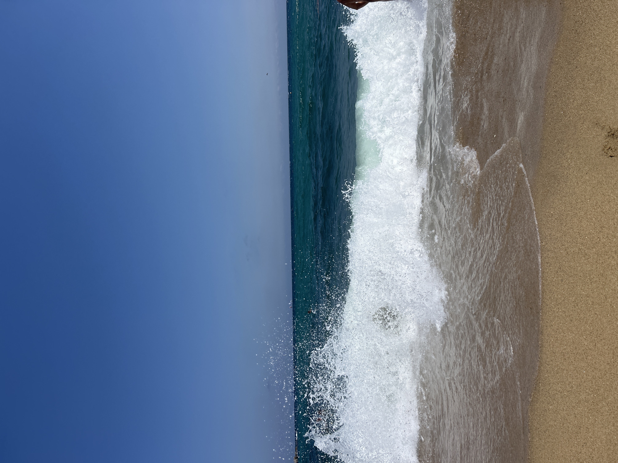The image depicts a serene beach scene with a clear blue sky and turquoise ocean water. A wave is captured mid-crash, with white foam and spray dominating the foreground. The sandy beach is visible at the bottom of the frame, with gentle waves lapping the shore. In the distance, the deep blue ocean stretches towards the horizon, with a few distant figures visible in the water, suggesting swimming or other water activities. The overall atmosphere is one of a bright, sunny day at a popular beach, likely in Barcelona, Spain, given its coastal location and vibrant Mediterranean feel.
