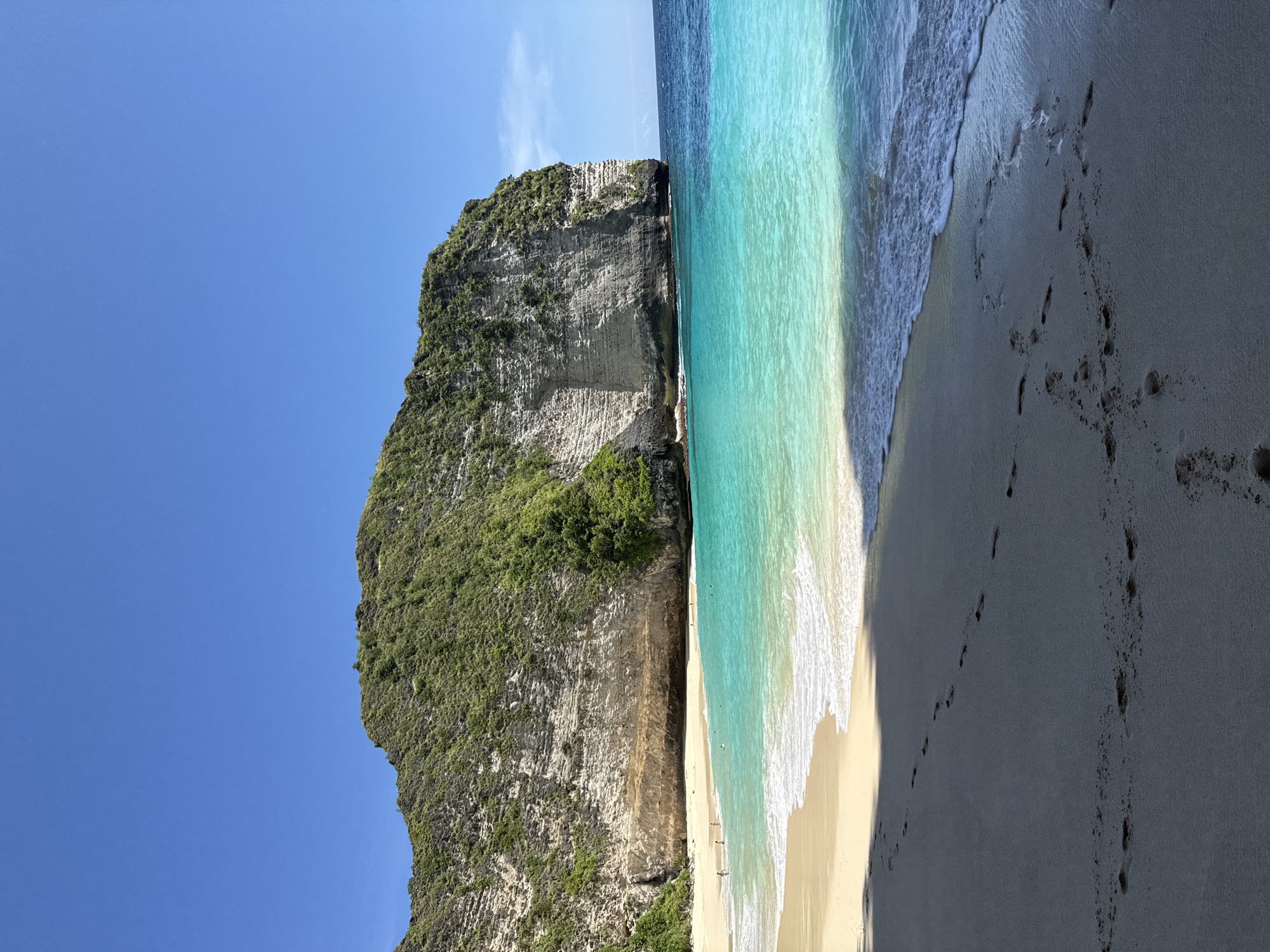 The image shows a scenic view of Kelingking Beach, located in Bunga Mekar, Nusa Penida, Indonesia. A large, verdant cliff dominates the left side of the image, its textured rock face partially covered in vegetation. Adjacent to the cliff is a narrow sandy beach, where a few people can be seen. The turquoise water meets the beach with gentle waves. Footprints are visible on the darker sand in the foreground. A clear blue sky completes the backdrop.