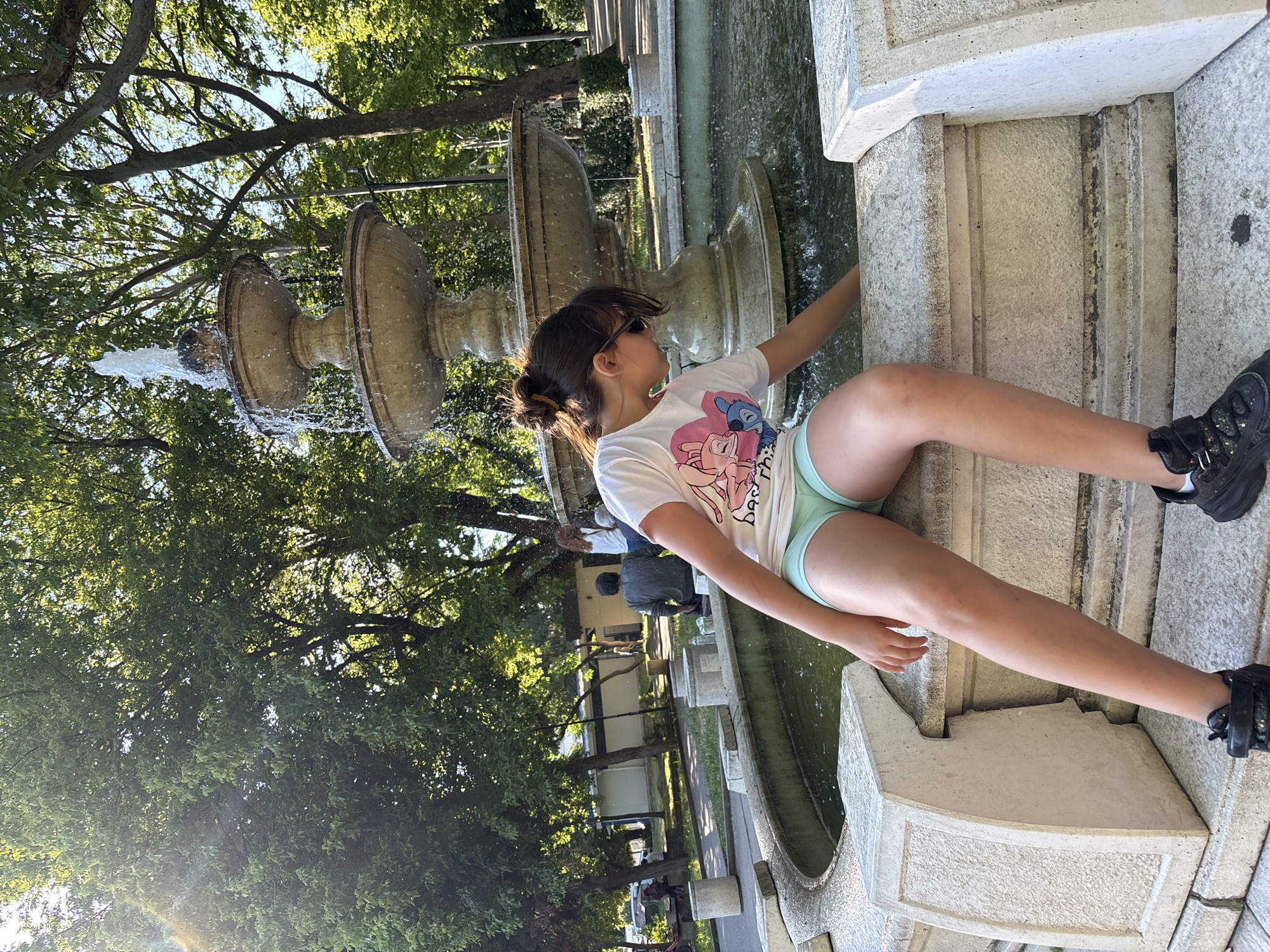 A young girl with dark hair pulled into a bun and sunglasses perched on her head is sitting on the edge of a stone fountain in a park. She is wearing a white t-shirt with graphics of cartoon characters and light blue shorts. Her right hand is reaching into the water. The fountain is multi-tiered and made of light-colored stone, with water spraying from the top. Behind the fountain, there are lush green trees providing shade, and a clear blue sky is visible. In the background, other people can be seen walking and sitting in the park, and there are some buildings. The scene appears to be a sunny day in a public park. This fountain is located in the city of Shumen, Bulgaria.