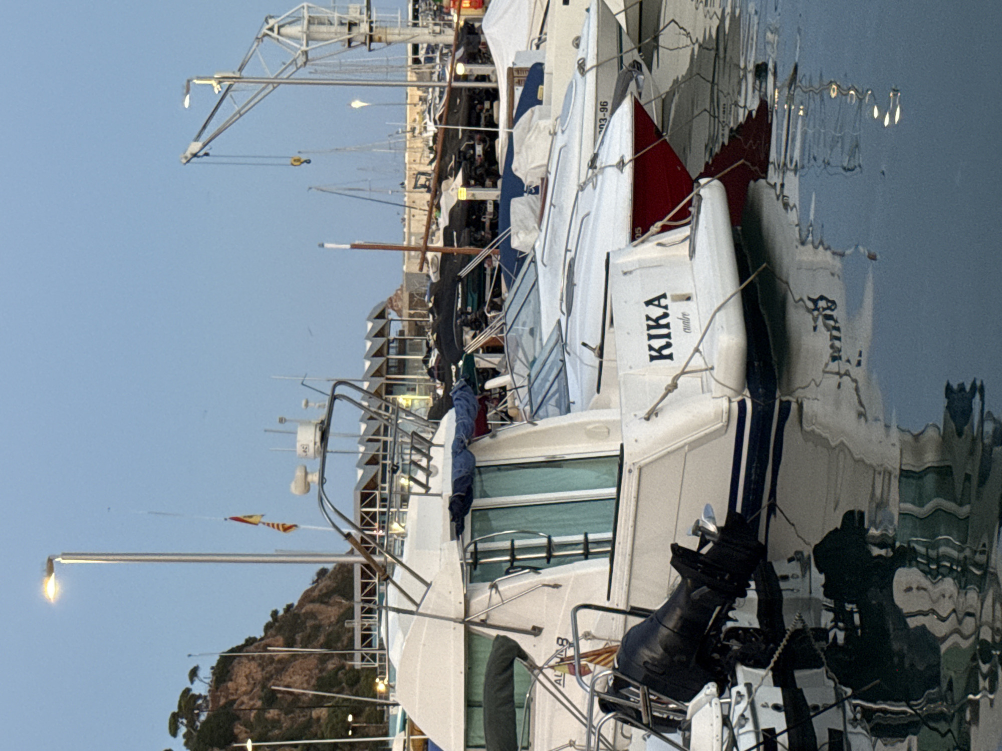 The image depicts a harbor scene in Blanes, Spain, likely in the early morning or late evening given the soft lighting and clear sky. The foreground features a white motorboat named "KIKA," with its name clearly visible on the stern. The boat has a black outboard motor and a ladder leading to the deck. The boat is docked alongside other vessels.
In the background, a marina with numerous boats of various sizes is visible. Several masts suggest sailboats are present. A large crane stands prominently on the right side of the image, indicating ongoing port activity or maintenance. In the distance, a rocky hillside covered with trees and buildings is visible, characteristic of the coastal landscape of Blanes. A streetlamp illuminates the scene. The water in the harbor is calm, reflecting the boats and the sky. No people are visible in the image. The overall impression is one of a quiet, operational harbor.