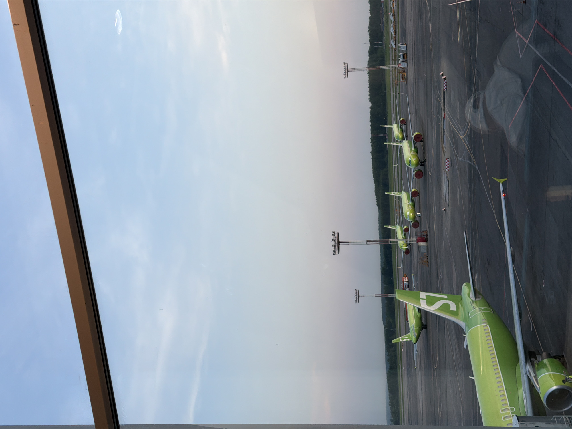 The image depicts an airport scene, likely at Domodedovo International Airport near Domodedovo, Russia, viewed through a window. Several green airplanes are parked on the tarmac, with tail fins visible. The airport environment includes ground markings, lighting structures, and a tree line in the background. The time of day appears to be daytime. The viewpoint is from inside a building, indicated by the window frame visible in the image's edge.