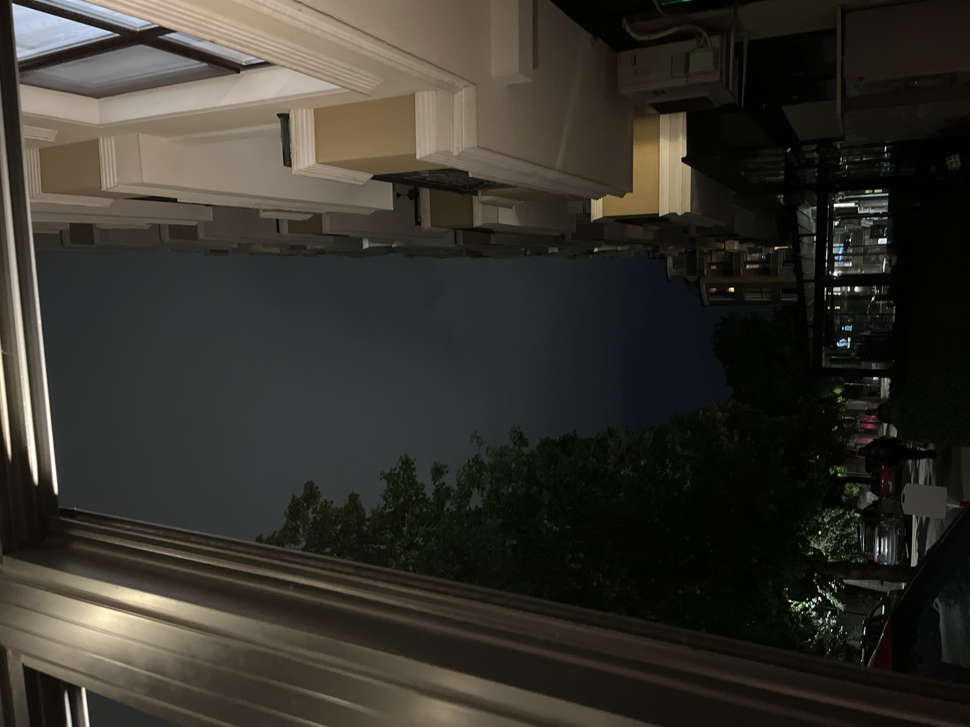 The image captures a street scene at night in Plovdiv, Bulgaria, likely near the Kapana creative district, given the architectural style of the buildings. The perspective is from a covered walkway or balcony, looking out towards a street lined with trees and buildings.
On the left side of the frame, a large, fluted column with a metallic finish dominates the foreground, partially obscuring the view. To its right, a dense, dark green tree with visible foliage is illuminated from below by streetlights, casting a dappled pattern on the ground and surrounding area.
The street itself is visible, with several cars parked along the side. One car, a silver SUV, is particularly noticeable. Further down the street, two people are walking, one of whom is a man in dark clothing. A red car is also visible parked on the street.
On the right side of the image, a modern apartment building with light-colored facades and balconies rises several stories. The building features decorative white trim around its windows and balconies. An outdoor air conditioning unit is mounted on the side of the building. Below the apartment building, at street level, are shopfronts with illuminated interiors, suggesting commercial activity. One of these appears to be a cafe or restaurant with glass windows and doors. In the distance, another building with lit windows can be seen.
The sky is dark, indicating it is nighttime, with a muted, overcast quality, suggesting possible fog or heavy cloud cover. The overall ambiance is urban and somewhat atmospheric, with the interplay of artificial lighting and natural elements creating a sense of depth and shadow.
