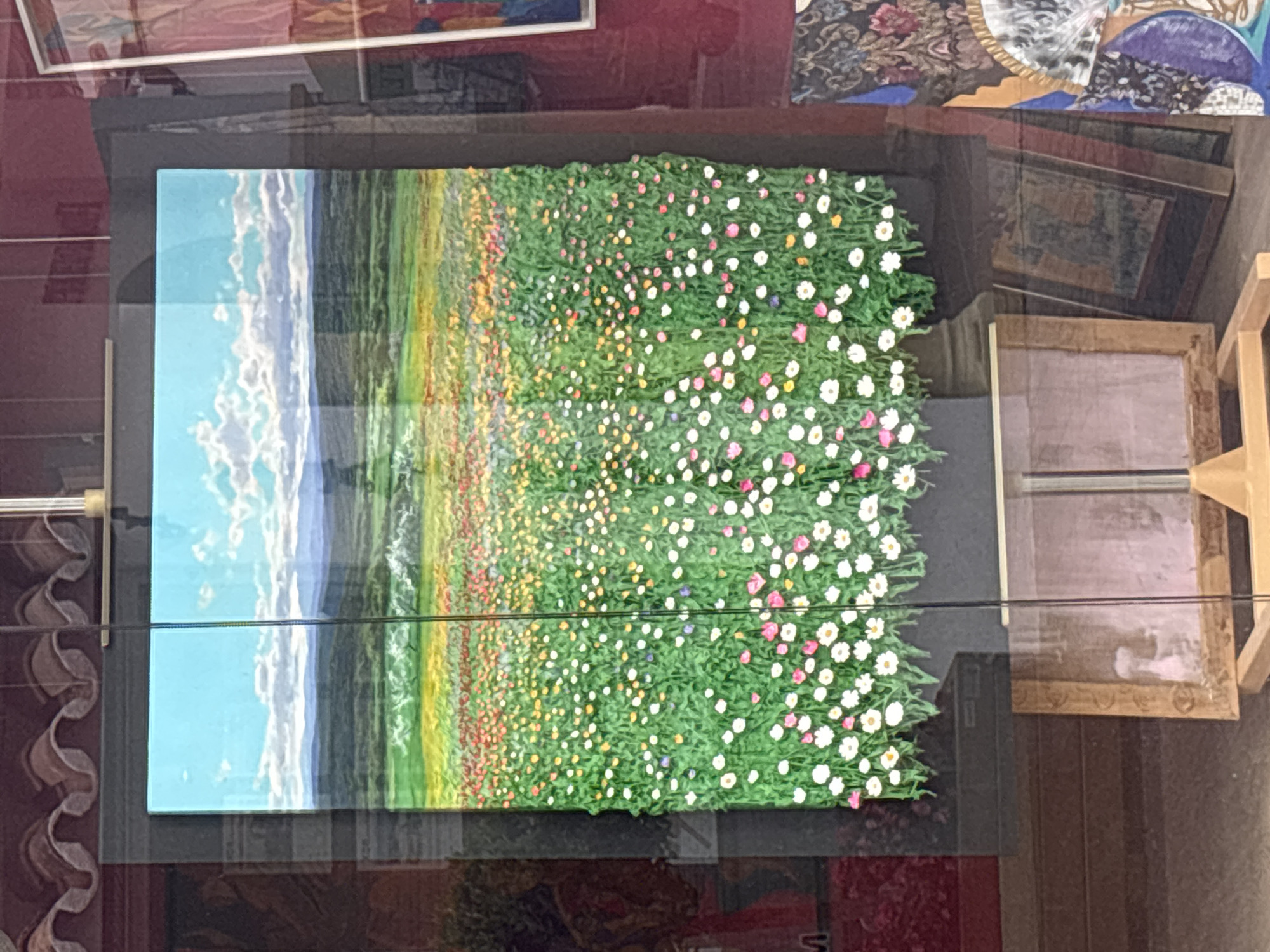 The image shows a painting of a vibrant wildflower meadow with a vast sky and distant mountains. The painting is displayed in a shop window, with reflections of the street and other objects visible on the glass. In the foreground, there are other framed artworks leaning against the wall.
The painting itself is a landscape featuring a dense field of flowers in various colors, including white, pink, yellow, and red. The flowers appear to be raised from the canvas, giving the painting a textured, almost three-dimensional quality. The meadow transitions to a green field, then to a distant range of mountains under a bright blue sky with fluffy white clouds.
The shop window has a dark frame and is partially obscured by a vertical support bar. Reflections on the glass show the exterior environment, including what appears to be a red building facade and hints of other shop displays. Below the painting, several framed pictures are visible, some leaning against each other, and one standing upright on a wooden easel. The floor of the shop appears to be tiled or paved.
The image was taken in Marbella, Spain, a city known for its affluent resorts and charming old town. While the specific location within Marbella is not provided, the scene suggests a typical boutique or art gallery setting commonly found in the city.