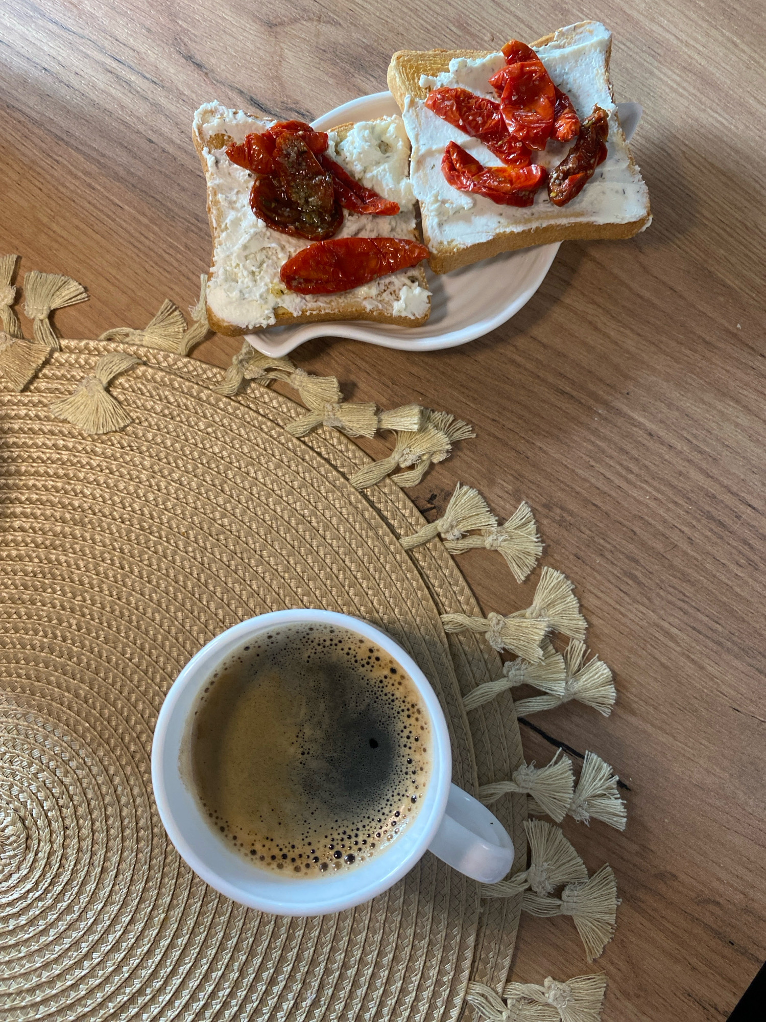 A high-angle shot captures a breakfast scene featuring two pieces of toast topped with cream cheese and sun-dried tomatoes, arranged on a white plate. Adjacent to the plate, on a circular woven placemat, sits a white mug filled with dark coffee. The placemat, adorned with cream-colored tassels, rests on a wooden surface. The composition is minimalist, focusing on the food and drink. The location of Zarechny, Russia is not depicted or inferable from the image.