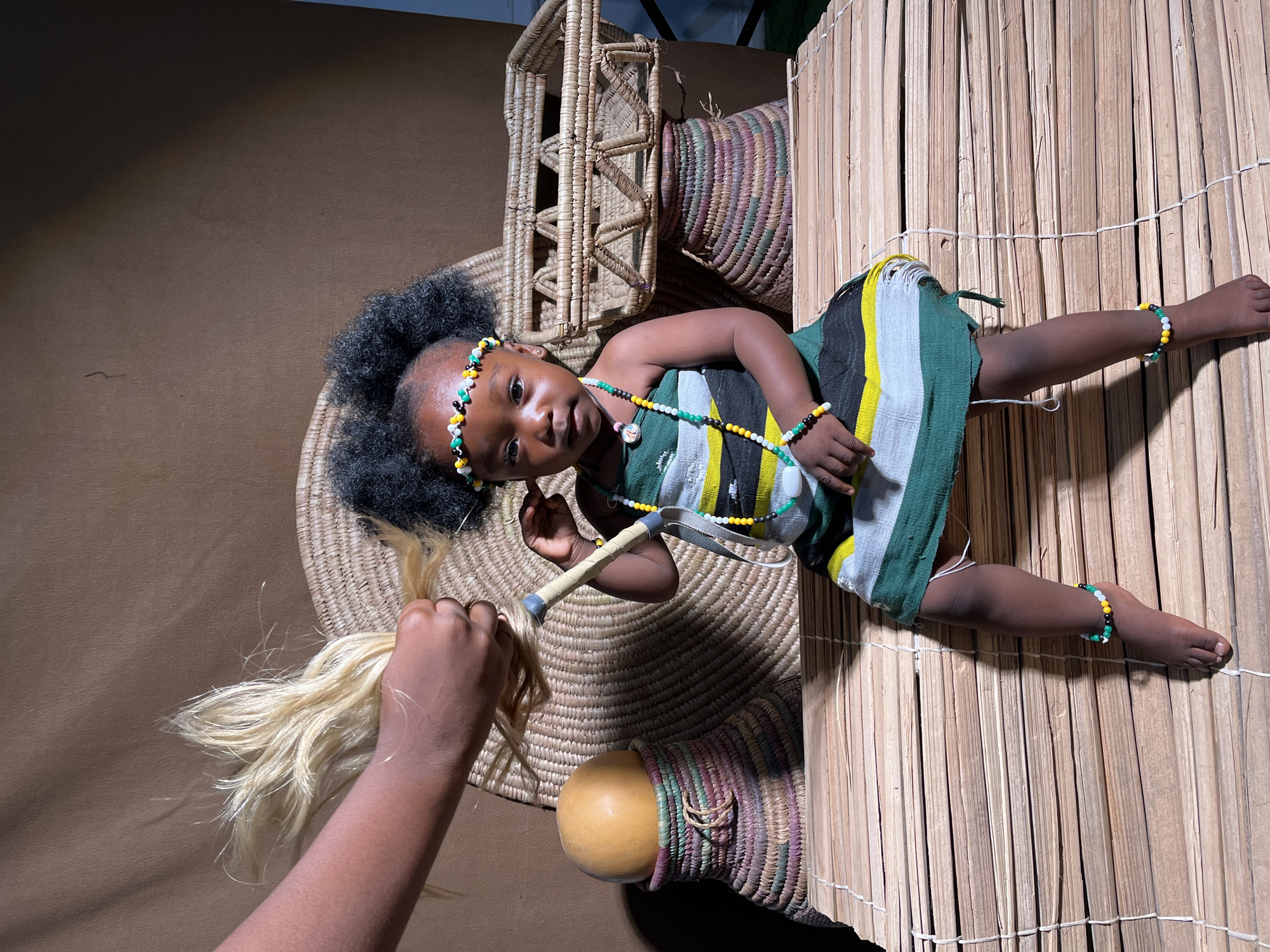 A child is lying down, slightly angled towards the camera. The child has dark skin, dark curly hair adorned with a beaded headband, and is wearing a dress with green, white, and black stripes and beaded jewelry. The child is holding a tool. A hand holding blonde strands of hair is visible in the lower left. The background features woven baskets, a gourd, and a bamboo-like structure. The location is in Barakin Akawo, Nigeria.
