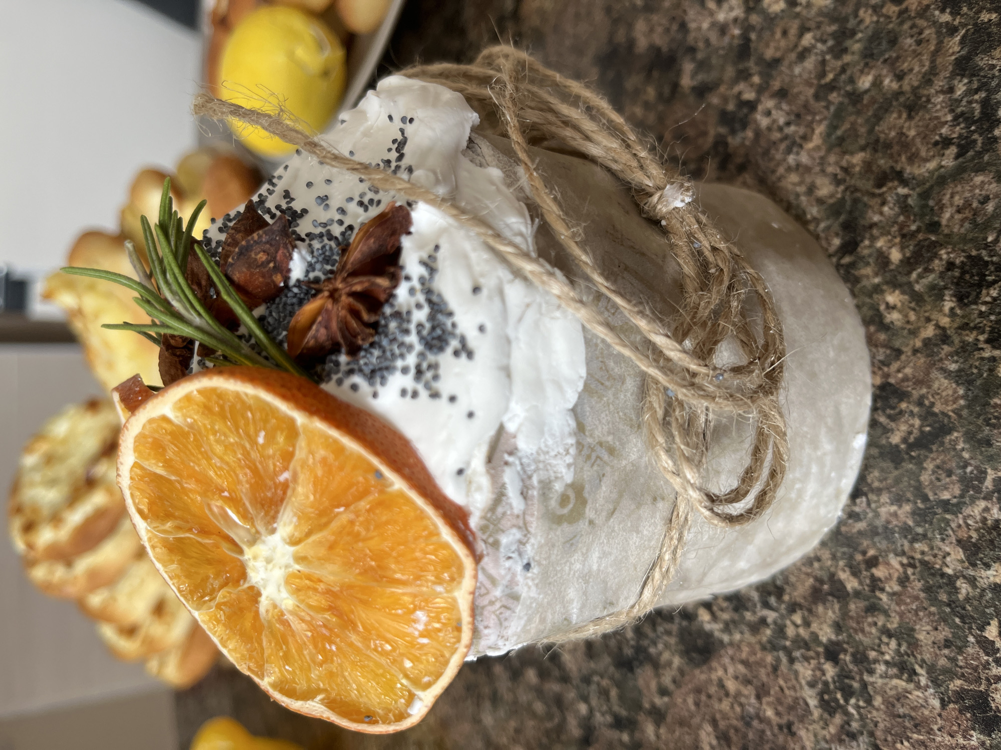 A top-down shot focuses on a cylindrical cheese, decorated and tied with twine. The cheese is covered in a white cream, sprinkled with poppy seeds. Garnish includes a dried orange slice, rosemary sprig, and dried star anise. The cheese sits on a brown countertop. A platter with bread and lemons is visible in the background. The location cannot be determined based on the image.