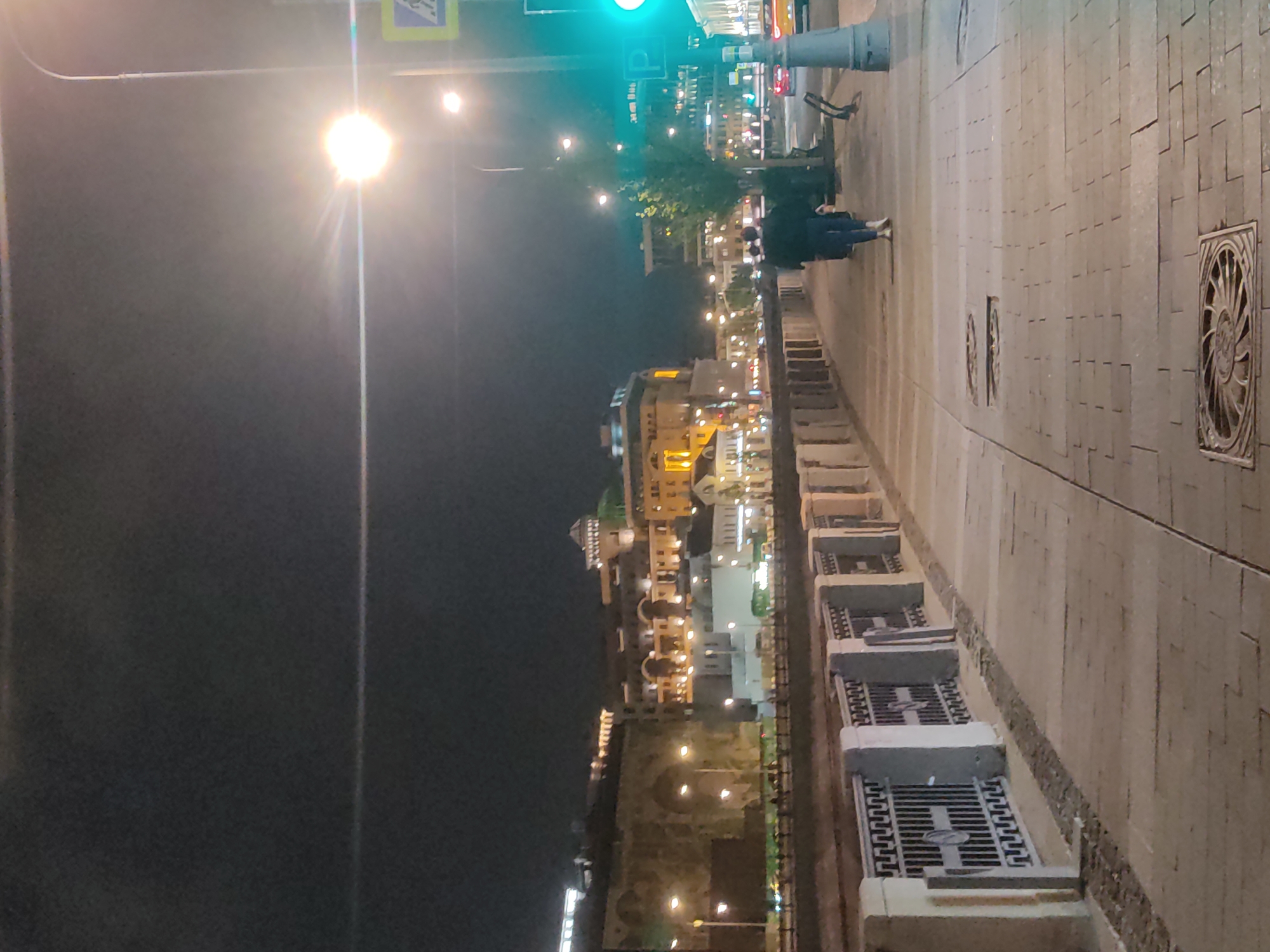 The image shows a nighttime scene in Moscow, Russia, likely along the Moskva River. The view includes a sidewalk with patterned stone paving, and the riverbank adorned with decorative fencing and stone ledges. In the distance, illuminated buildings with architectural detail, including a distinctive yellow building with lights, are visible. Street lights line the riverbank, casting a glow on the surroundings. Two people are walking along the sidewalk. A street light and traffic signal are also visible.