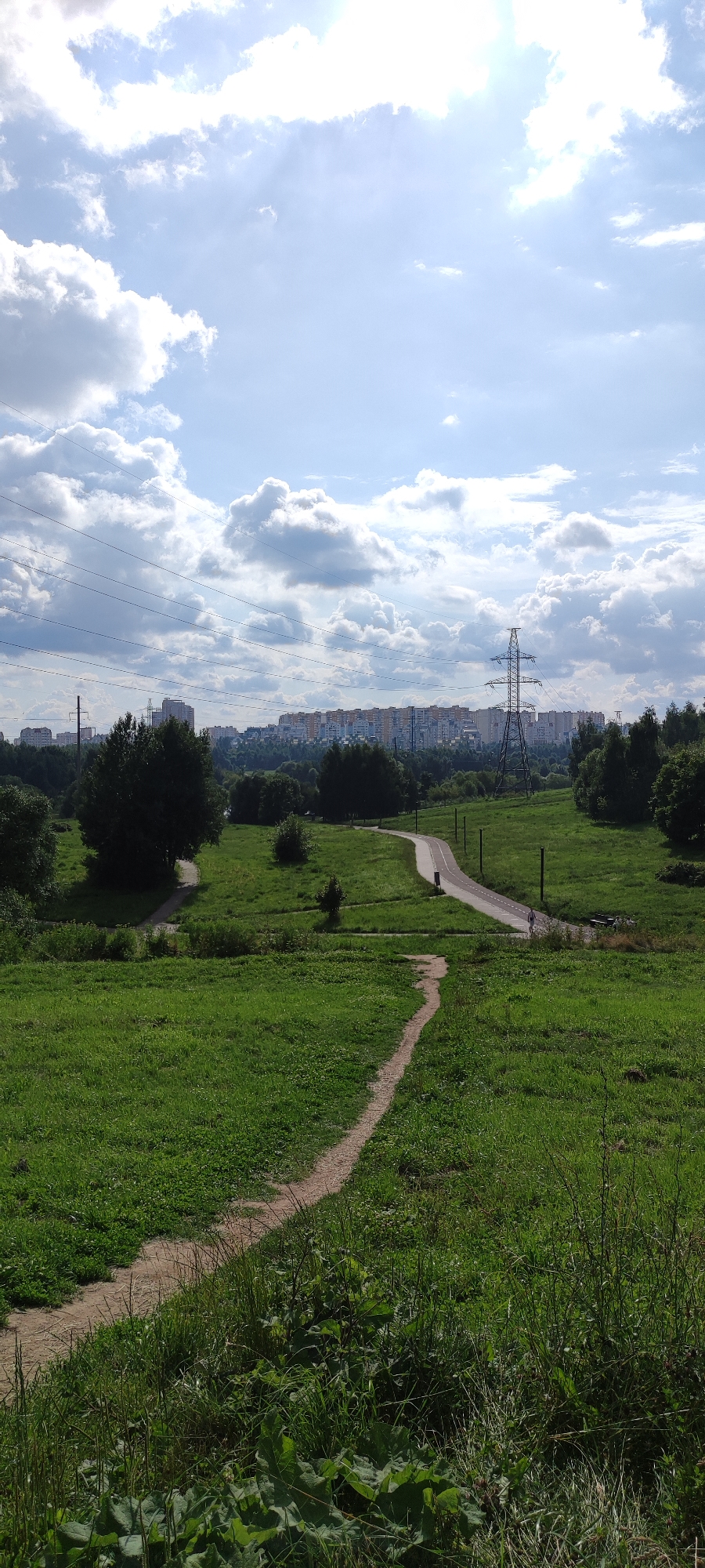 The image depicts a panoramic view of a suburban landscape in Khimki, Russia, under a partially cloudy sky with sunlight filtering through. In the foreground, a dirt path winds its way through a lush green meadow dotted with trees and bushes. The path curves to the right, leading towards a paved road with a cycling lane. In the middle ground, a cluster of trees forms a dark green line against the horizon. Beyond the trees, a vast urban panorama unfolds, characterized by rows of multi-story apartment buildings, suggesting a densely populated residential area. A prominent power pylon stands tall against the sky, with electrical wires extending across the frame, signifying the infrastructure of the city. In the distance, the city stretches across the horizon, merging with the sky under the bright sunlight. The scene suggests a typical day in a Russian suburb, with a blend of natural landscapes and urban development.