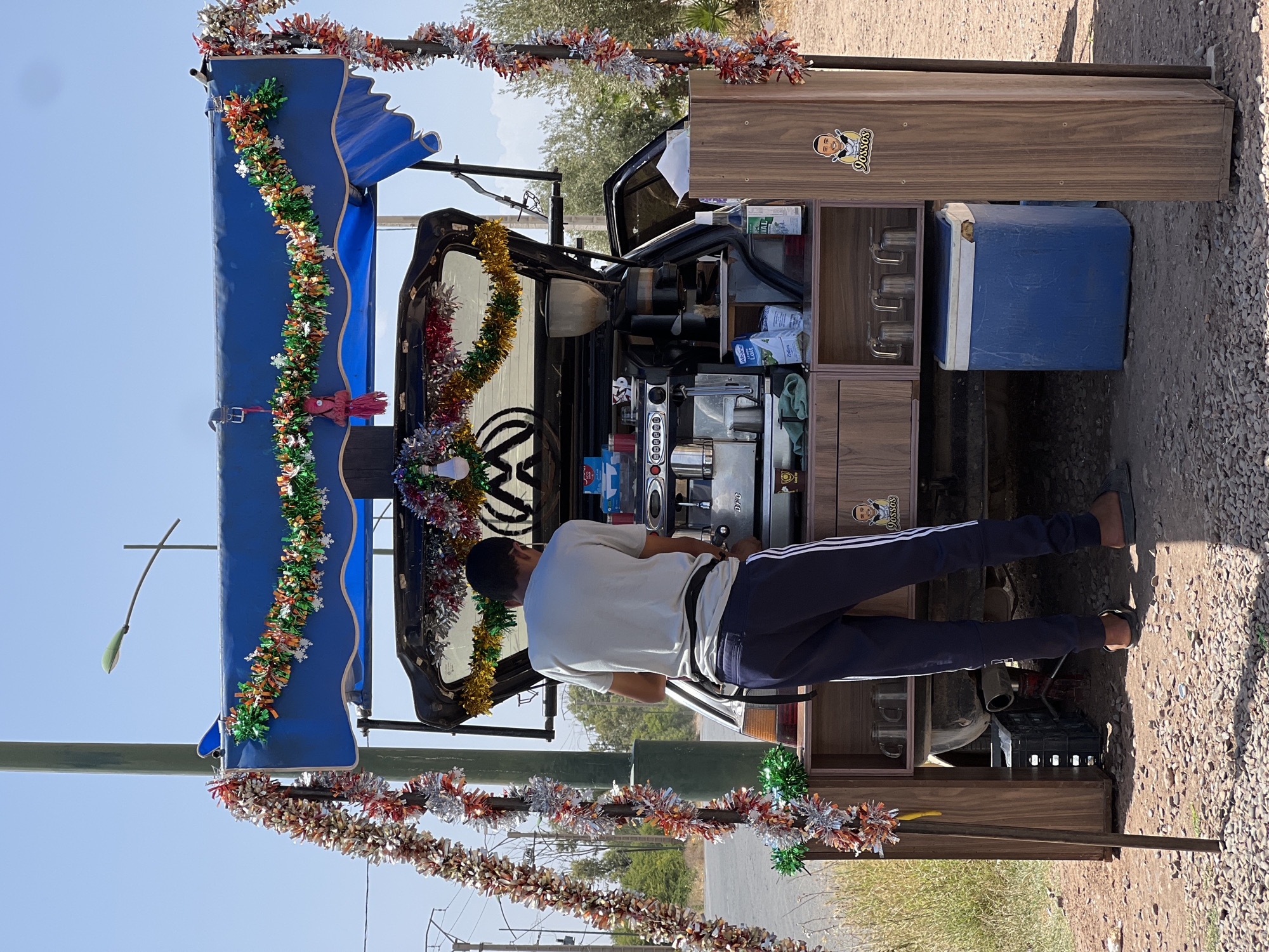 A man operates a mobile coffee stand constructed from the rear of a Volkswagen vehicle, Saâda, Morocco. The vehicle's back end is fitted with a wooden cabinet containing coffee-making equipment, including an espresso machine, cups, and various beverages. A blue cooler is positioned beside the cabinet. The vehicle's roof is partially covered by a blue awning. The stand is decorated with colorful metallic garlands. The man, wearing a white t-shirt and blue track pants, stands behind the cabinet operating the espresso machine. The scene is set outdoors on a dirt road. The weather appears sunny and clear. The mobile coffee stand is located on the side of a road in a rural setting with trees and vegetation visible in the background.