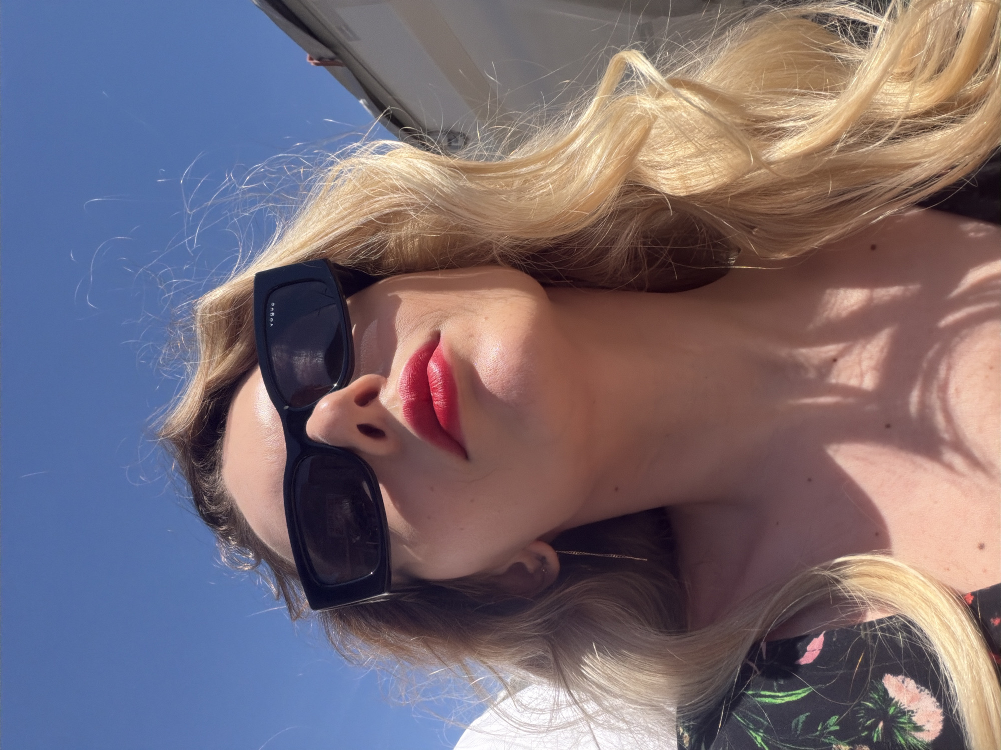 A woman with long blonde wavy hair is seen from a low angle, with a clear blue sky partially visible in the background. She is wearing large, black sunglasses and bright red lipstick. The woman is wearing a floral print top or dress, and gold earrings are visible. The top of a white structure is also visible in the top left corner of the frame. There is no location information, so I cannot determine if this is in Budva, Montenegro.