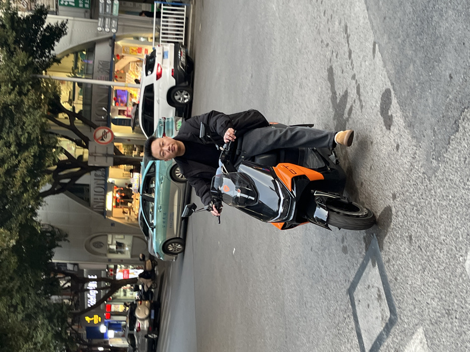 An adult East Asian male is seated on a black and orange "GT" model electric scooter on a paved city street in Jiangjin, Chongqing, China. He wears a dark puffer jacket over a dark undershirt, dark grey trousers, and light brown slip-on shoes, with both hands gripping the scooter's handlebars. His expression is neutral as he looks directly forward. The asphalt road surface beneath the scooter features lighter patches and dark stains, with a distinct square shadow cast near the front wheel. In the background, multi-story commercial buildings line the street, displaying illuminated storefronts, including one labeled "INHON(G)" and another featuring "足浴" (foot bath) signage. Several vehicles are visible, including a teal sedan identified as a Chongqing taxi and a white SUV. Green-foliaged trees, a white metal pedestrian barrier, and a red circular "no honking" traffic sign are also present. The scene is illuminated by a combination of ambient natural light and artificial street and store lighting, suggesting late afternoon or early evening conditions.