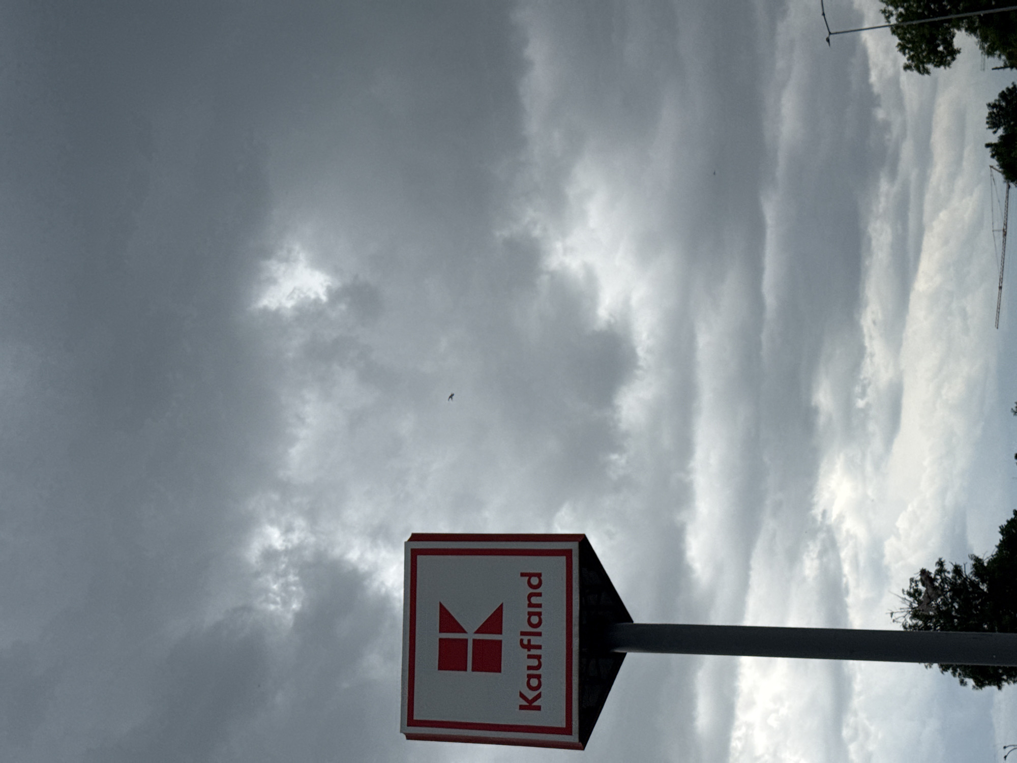 The image shows a Kaufland sign against a cloudy sky. The sign is red and white with the word "Kaufland" written vertically. The sky is mostly gray with patches of lighter clouds. The location is likely near a Kaufland store, potentially in the city of Burgas, Bulgaria. Trees and a crane are partially visible in the upper right corner of the image.