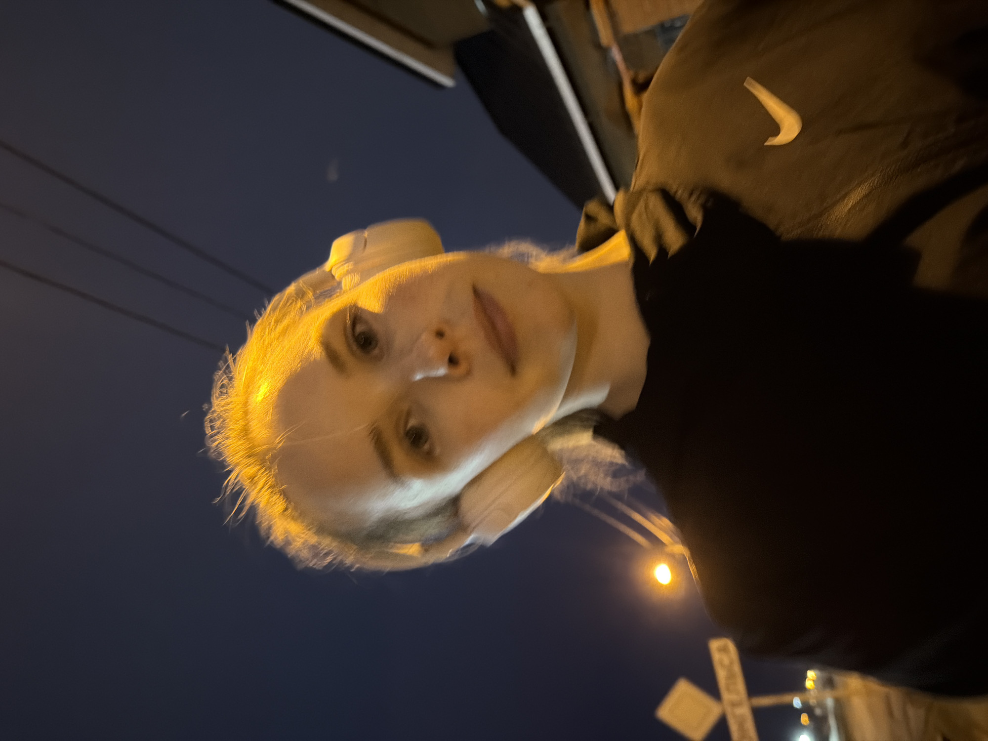 A woman with fair skin and blonde hair wearing white headphones and a black Nike jacket is standing near a railroad crossing. The environment appears to be at night in the city of Berezniki, Russia.