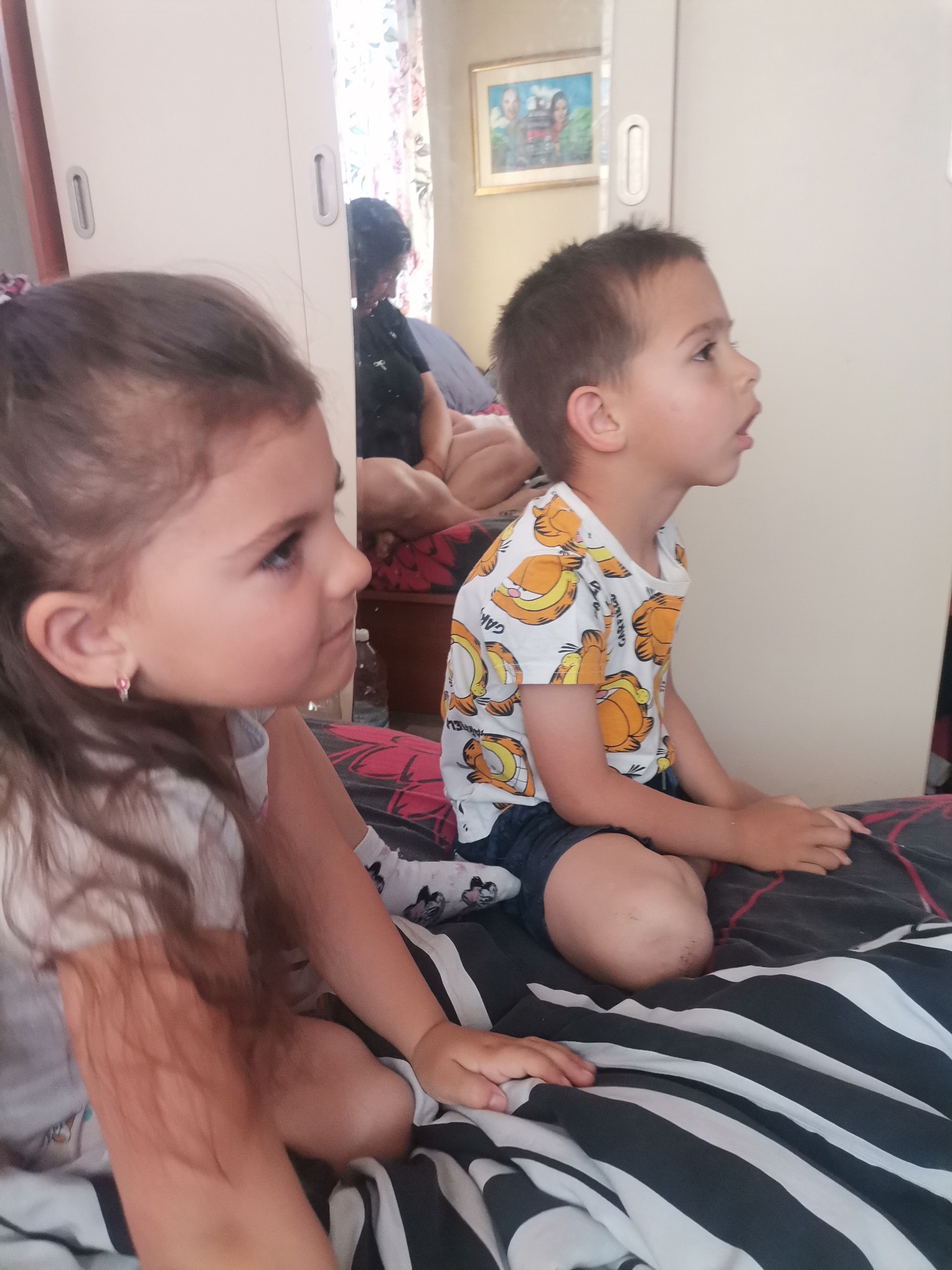 An indoor photograph captures two young children, a girl and a boy, sitting side-by-side on a bed, engrossed in watching something off-camera to their right. The girl, closer to the foreground, has long brown hair and wears a white t-shirt with a small pink earring visible on her left ear. Beside her, the boy wears a white t-shirt patterned with orange Garfield characters, with the text "GARFIELD" visible, and dark shorts.
They are seated on a bed, which is partially covered by a black and white striped duvet. Another dark, patterned fabric with red accents is visible beneath the boy. In the background, a light-colored wardrobe with sliding doors reflects parts of the room. Within this reflection, an adult with dark hair is visible, seated further back in the room, and a framed picture hanging on a wall can be seen. The picture has a golden frame and depicts two people. A water bottle is partially visible near the boy's leg. The setting appears to be a bedroom during the daytime, indicated by the ambient light. Both children exhibit focused and calm expressions, seemingly captivated by what they are viewing.