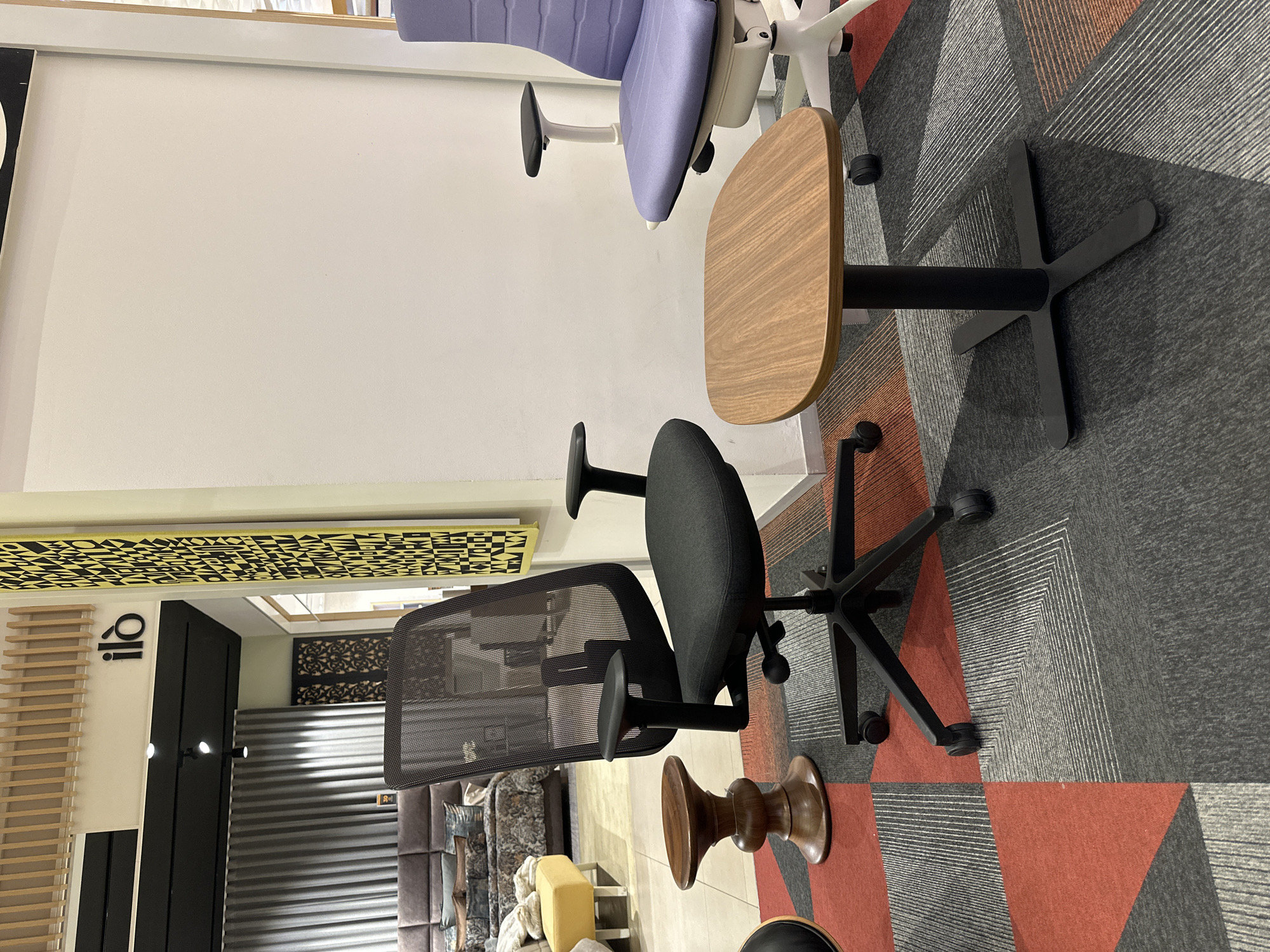 The image shows a display of office furniture inside a building. The furniture includes a purple and white ergonomic chair, a desk, and two black office chairs with mesh backs. The chairs are on a gray carpet with a red geometric pattern. The walls are white, and there is a sign with black lettering in the background. The location may be in Mexico City, Mexico.