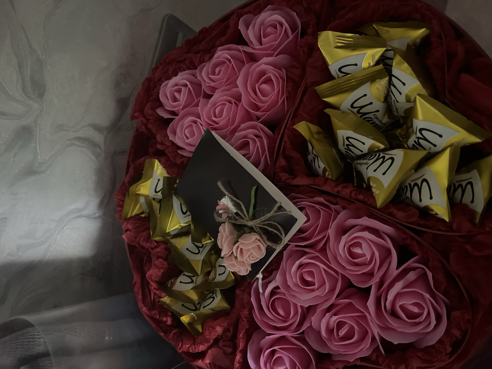 In Bohunove, Ukraine, a heart-shaped gift box is presented, filled with an assortment of items. The box is lined with red fabric, creating a vibrant backdrop. Within the arrangement, numerous pink, artificial roses are meticulously placed, their petals unfurled in full bloom. Interspersed among the roses are several individually wrapped chocolates, each adorned with a golden wrapper featuring black lettering. A small, dark-colored notebook rests on top of the chocolates and roses, decorated with a miniature bouquet of pink roses tied with twine. The surrounding environment is dimly lit, with a textured, marbled wall visible in the background, suggesting an indoor setting.