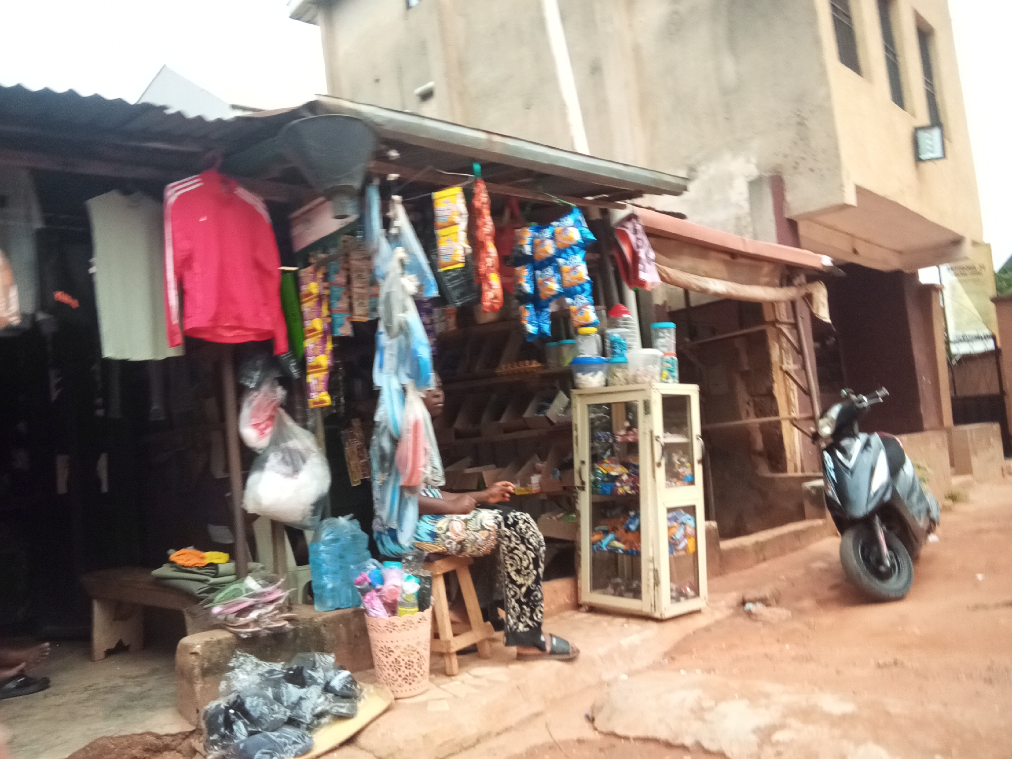In Oko Town, Nigeria, a modest roadside shop is depicted with a variety of goods on display. A woman, dressed in a blue and white patterned top and black and white patterned trousers, sits on a wooden stool in front of the shop, seemingly attending to customers or sorting items. To her right, a light-colored display cabinet showcases an assortment of packaged goods.
The shop's facade features a corrugated metal roof providing shelter. Various items, including clothing like a red jacket with white stripes, and numerous packets of snacks and other products, hang from hooks or are displayed on shelves within and outside the stall. A blue plastic bag, a basket filled with colorful items, and other merchandise are visible on the ground and on a low wooden bench. A dark-colored scooter is parked to the right of the shop. The ground is covered in reddish-brown earth, typical of the environment in Oko Town. The building behind the shop appears to be a multi-story structure with windows.
