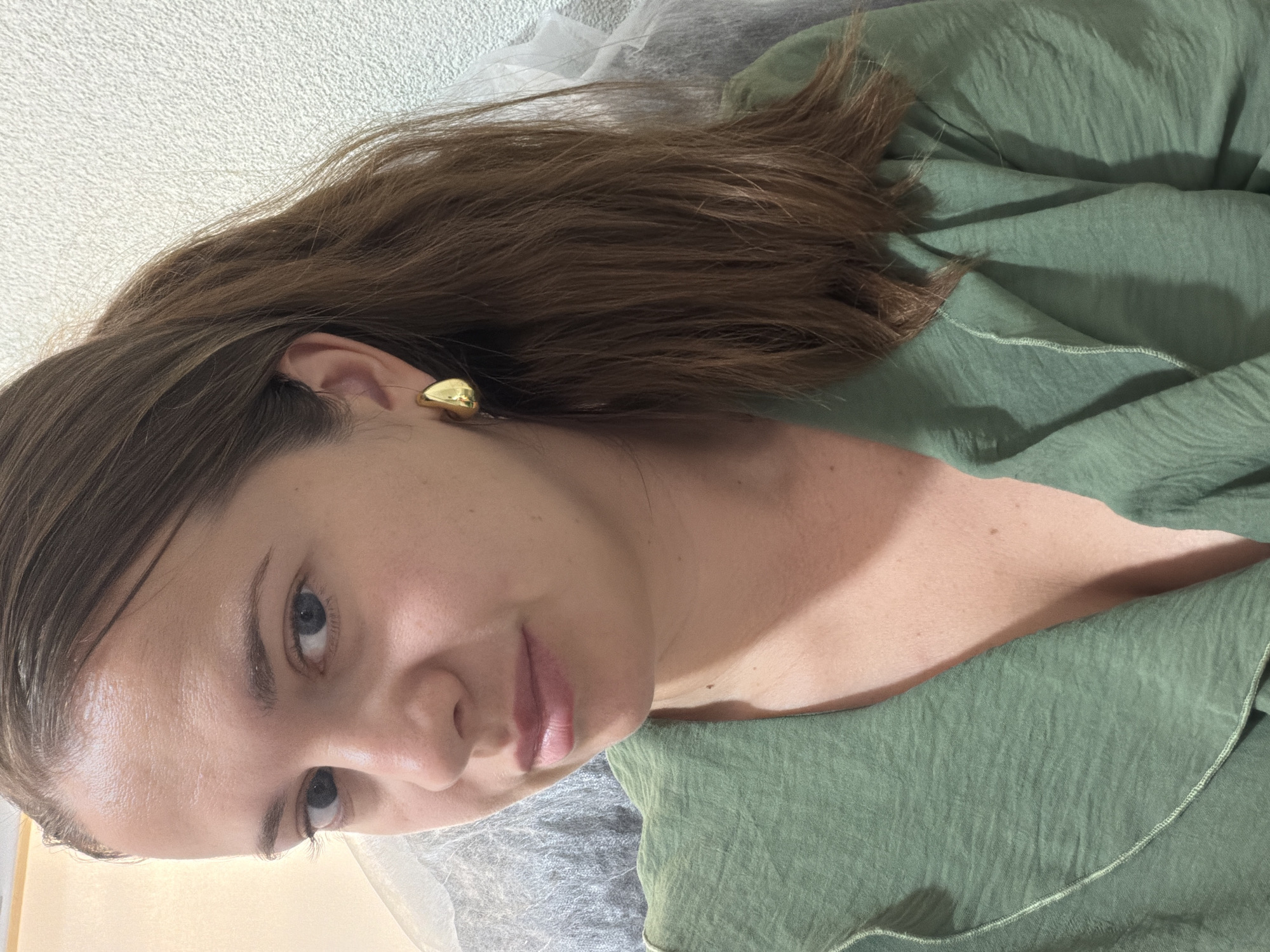 The image shows a medium shot of a woman with brown hair pulled back in a ponytail, wearing a green top and gold earrings. She has blue eyes and is looking towards the camera. The background is out of focus and appears to be an indoor setting with a textured wall. No location in the city of Sochi, Russia, is identifiable from the image.
