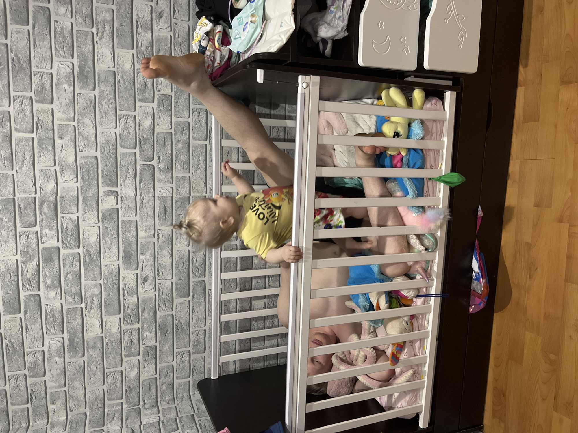 A man and a toddler are inside a white crib filled with toys. The man's head is closest to the bottom of the frame, and his legs extend upwards towards the top of the frame. The toddler is holding onto the side of the crib. The crib is part of a dark brown dresser. The location appears to be indoors, with a gray brick-patterned wallpaper on the left and a wooden floor on the right. The top of the dresser is cluttered with clothes and diapers. The location, Vesyoloye, Russia is not verifiable from the image.